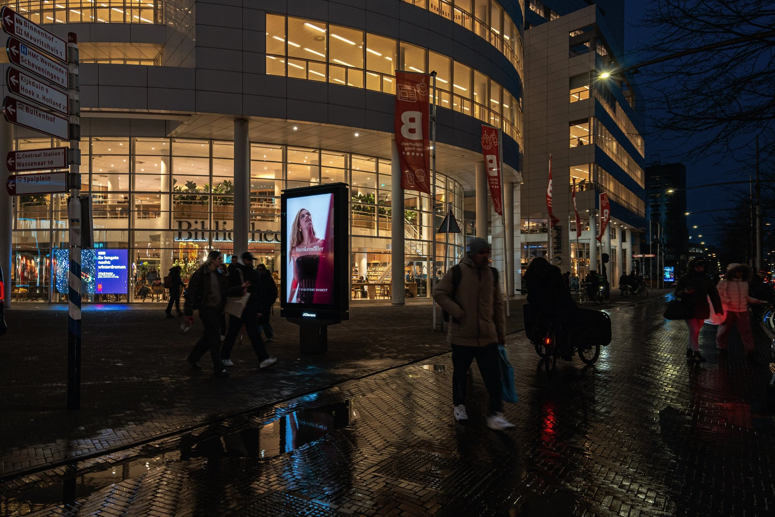 Stadshuis met mensen die voorbij lopen, verlichte gevels en digitale reclameborden in de avond.