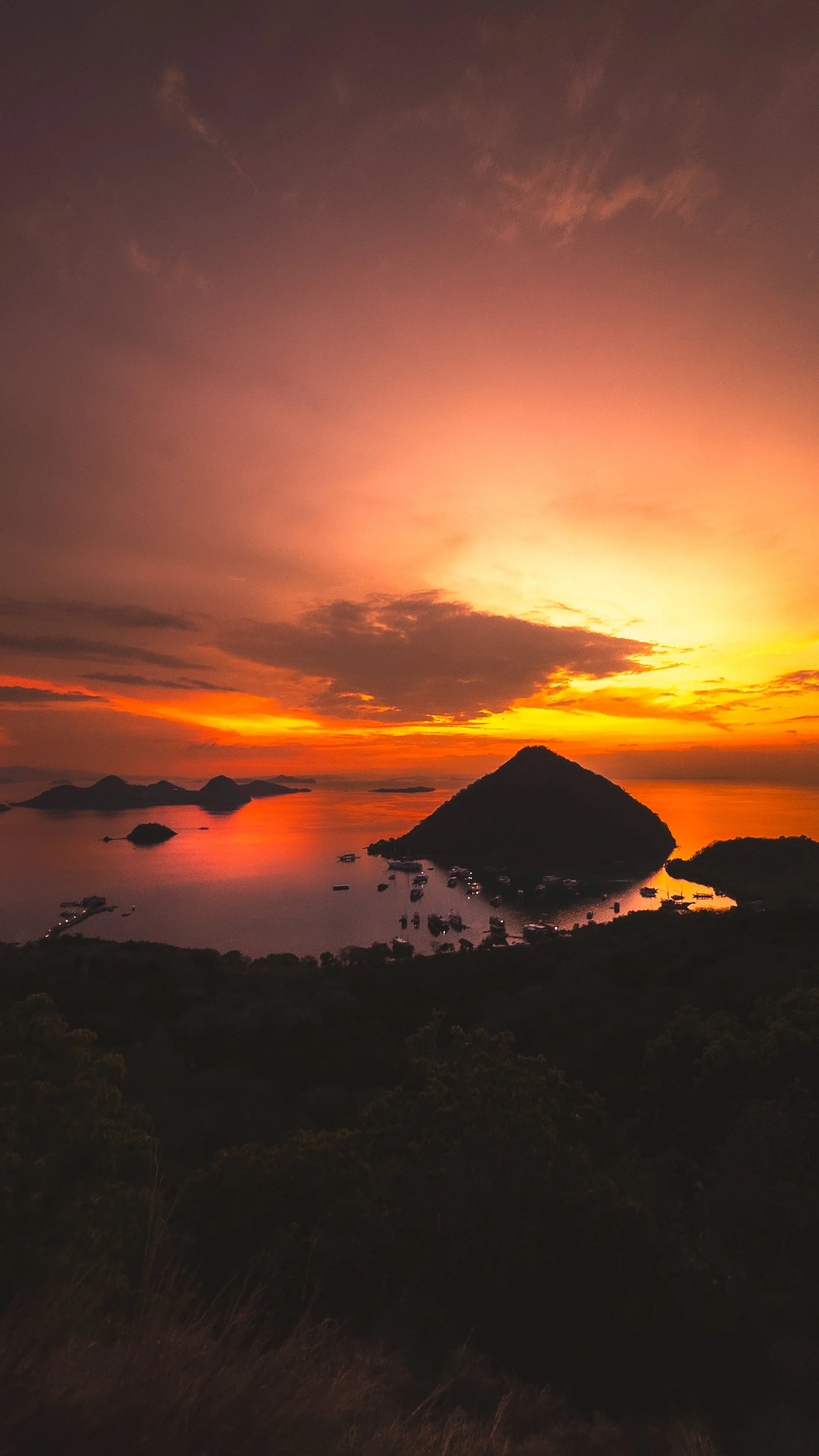 Uitzicht op een zonsondergang over een eiland met een berg, omgeven door water en kleine boten. De lucht is gekleurd in roze, oranje en geel.