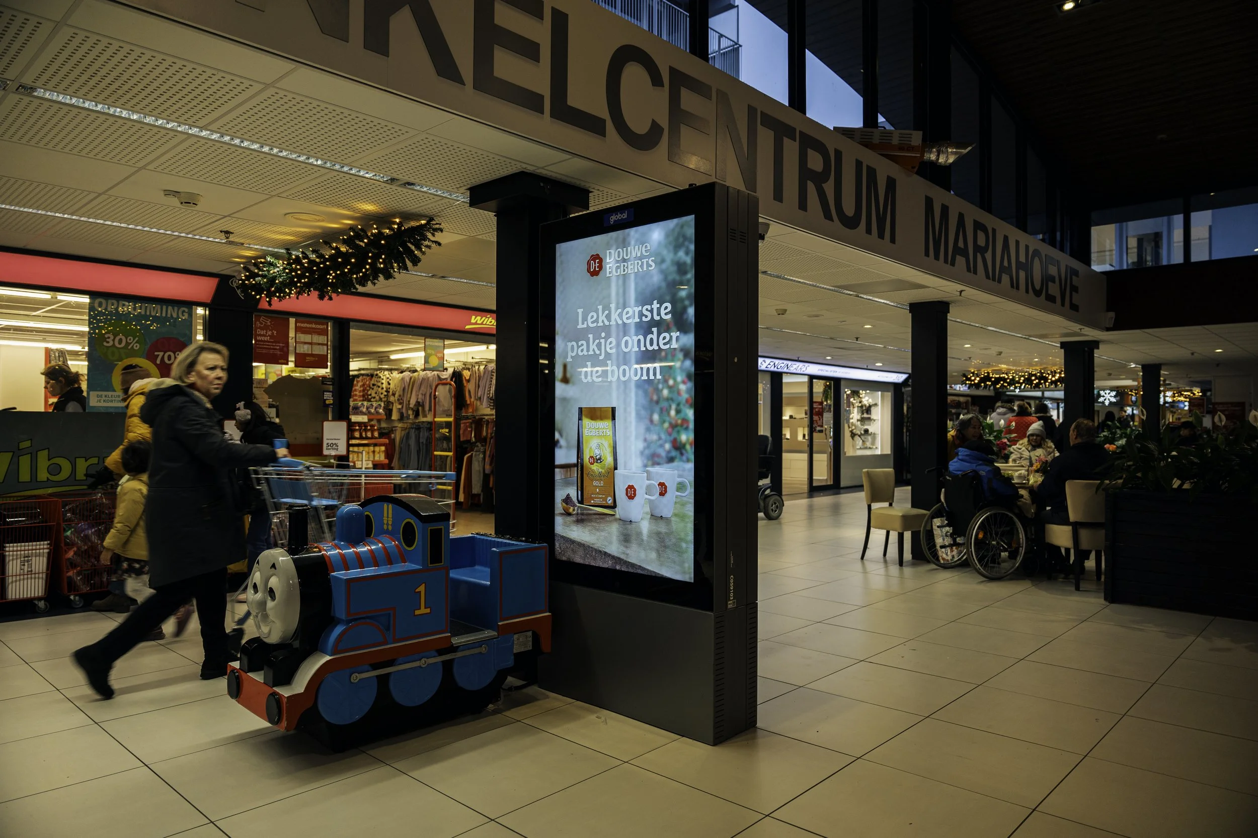 Binnen in een winkelcentrum met een kleine kindertrein en klanten die winkelen en aan een tafel zitten. Een digitale advertentiebord en winkels op de achtergrond, met decoraties voor de feestdagen.