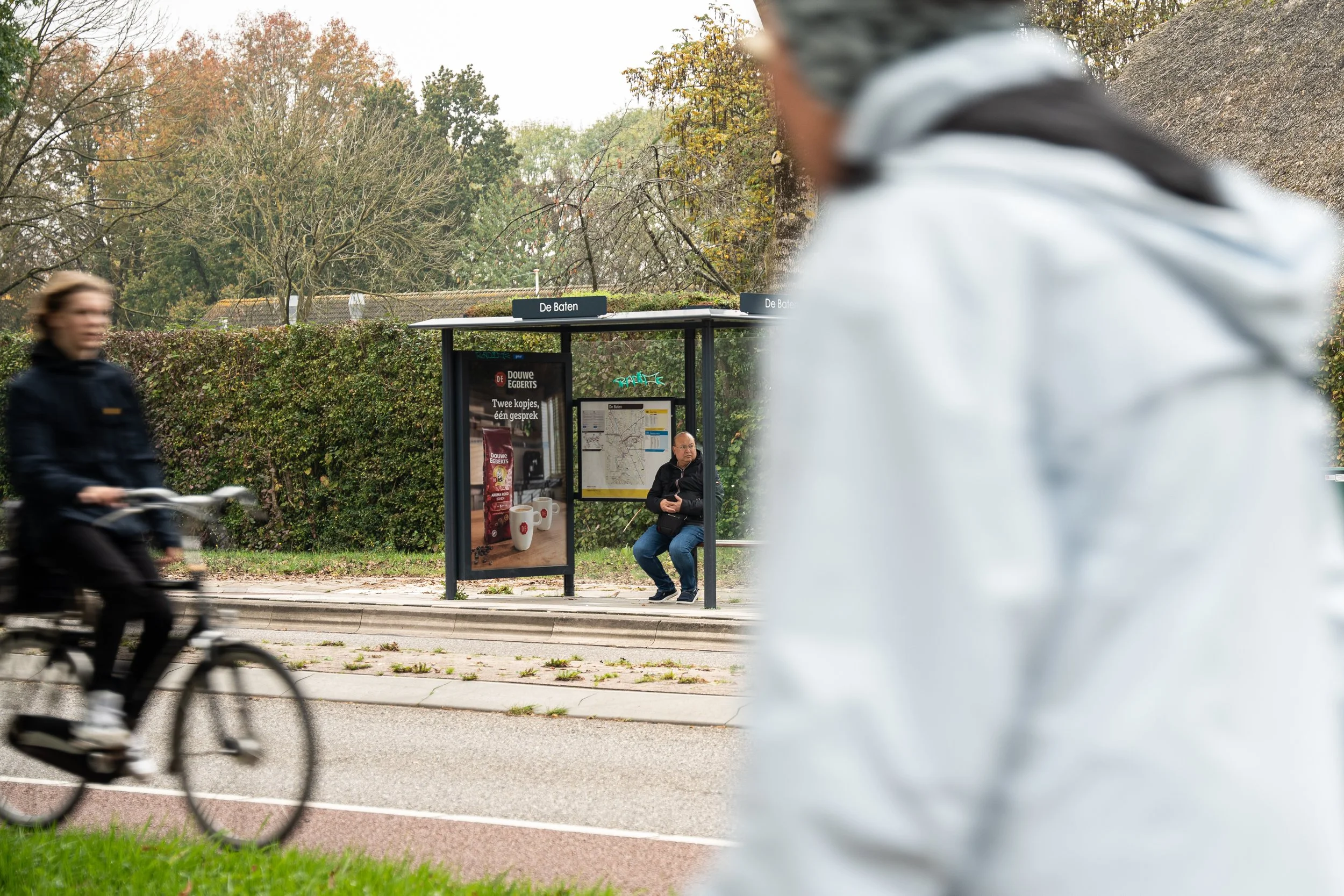 Man zit op een bushokje, terwijl een vrouw op de fiets passeert en een persoon met een witte jas op de voorgrond zichtbaar is.