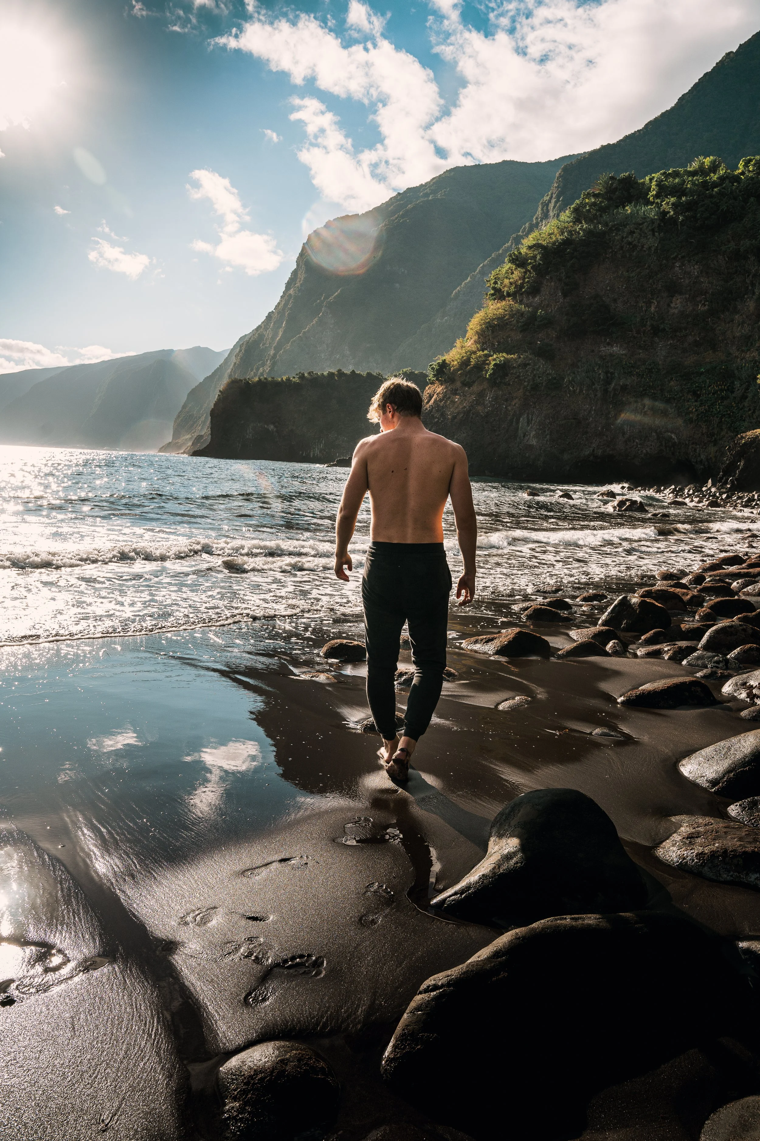 Man loopt langs de kust op een zwart strand met rotsen, omgeven door groene bergen en een blauwe lucht met wolken.