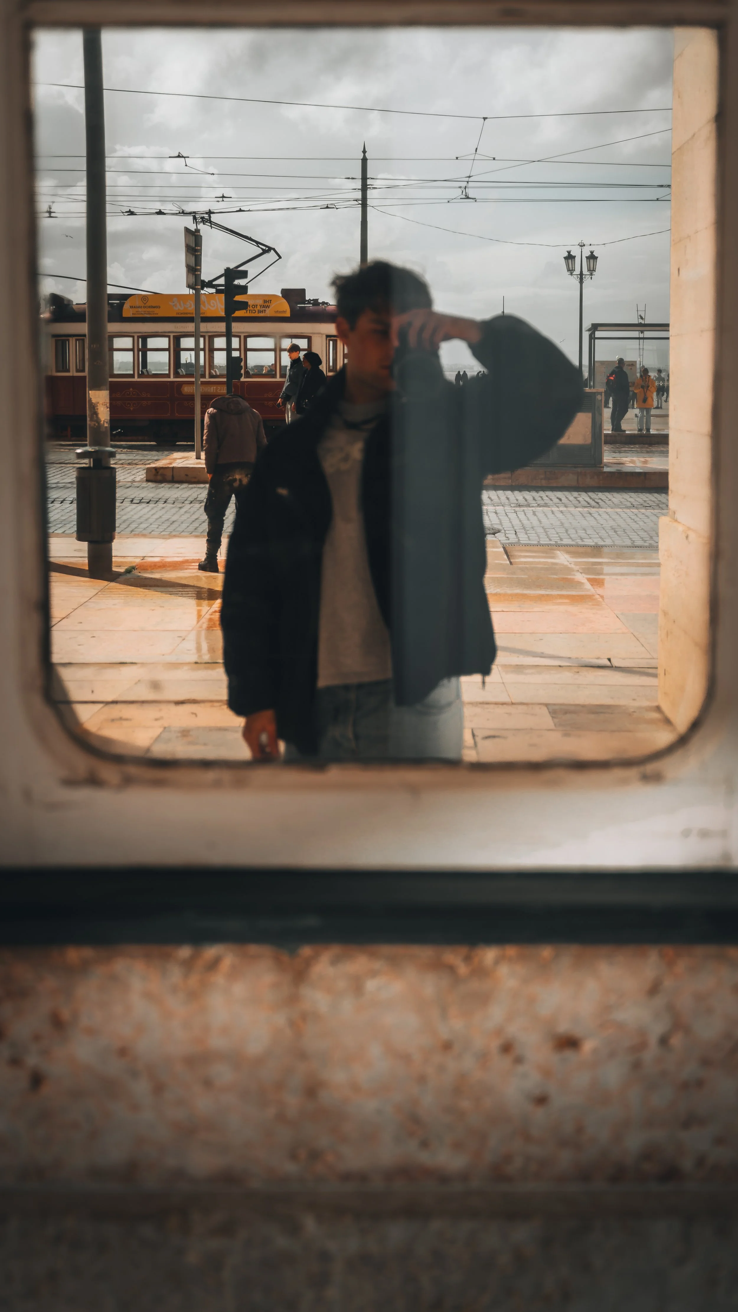 Reflectie van een jonge man in een raam met een tram en enkele wandelaars op het station op de achtergrond.
