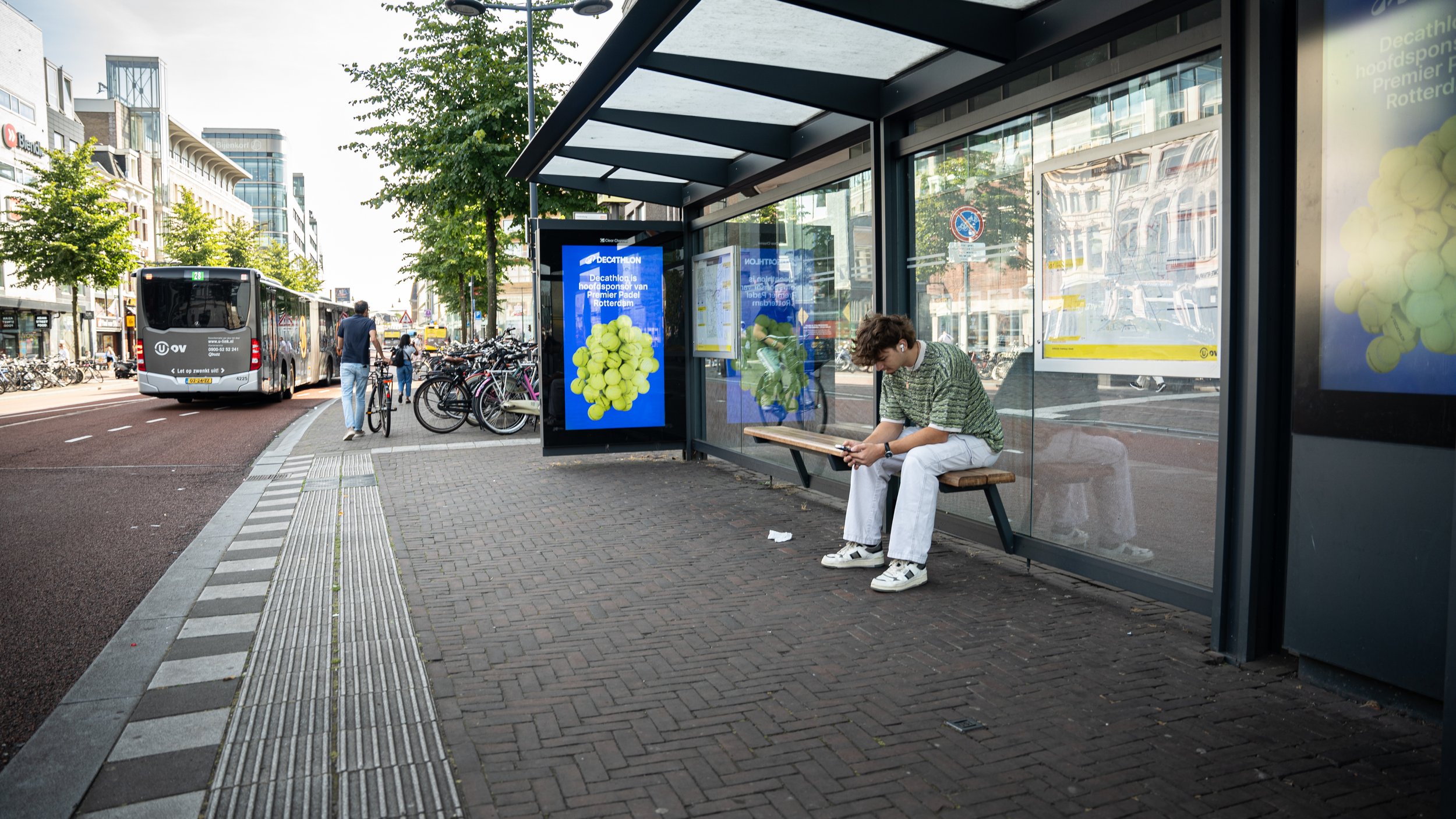 Een jonge persoon zit op een bankje bij een bushokje op een stadsplein, kijkt op een tablet of telefoon, en andere mensen lopen of fietsen in de straat.