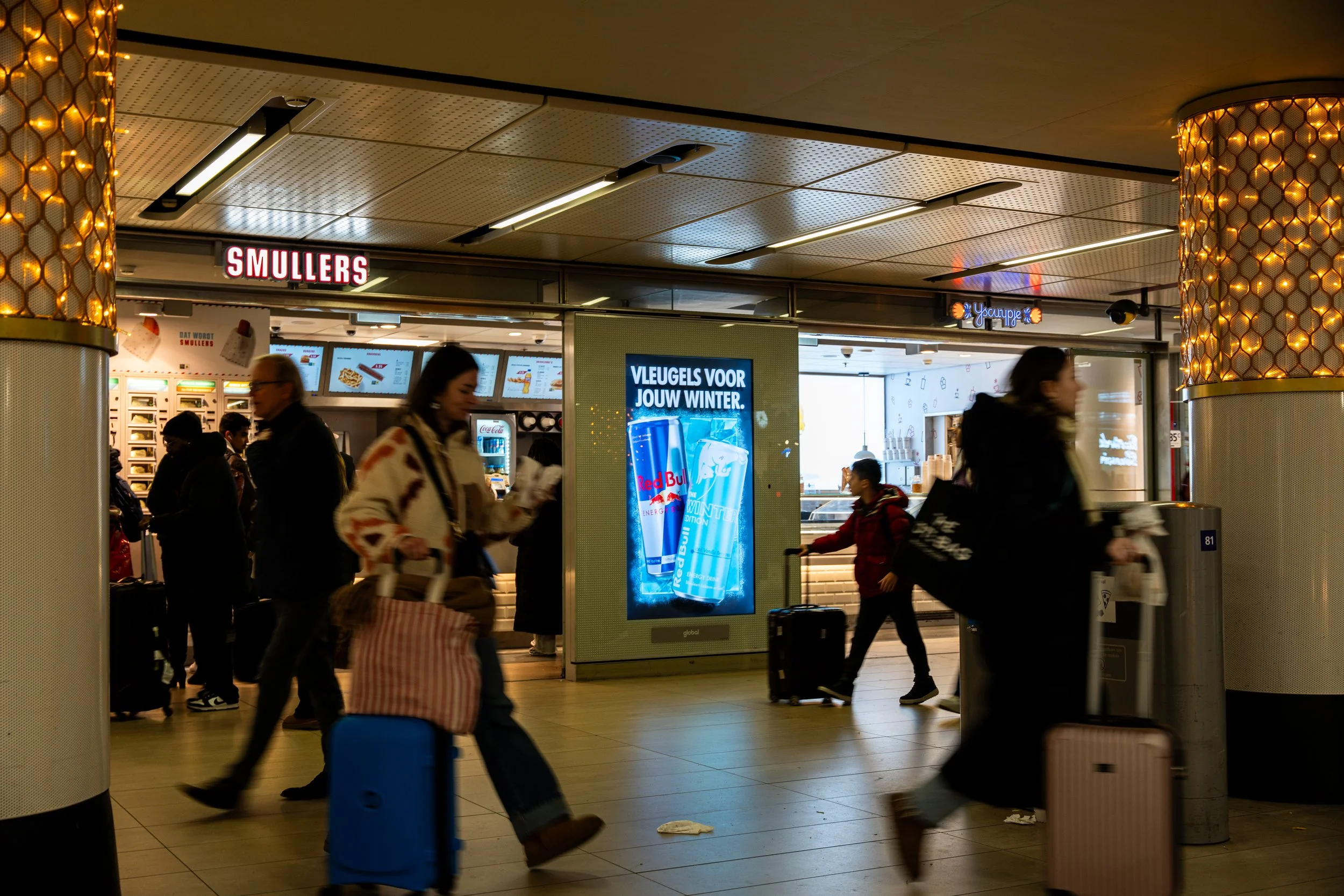 Slecht verlichte luchthaven met mensen die lopen met bagage, reclamebord voor Red Bull, snackbar genaamd Smullers, en een digitale advertentie in het midden.