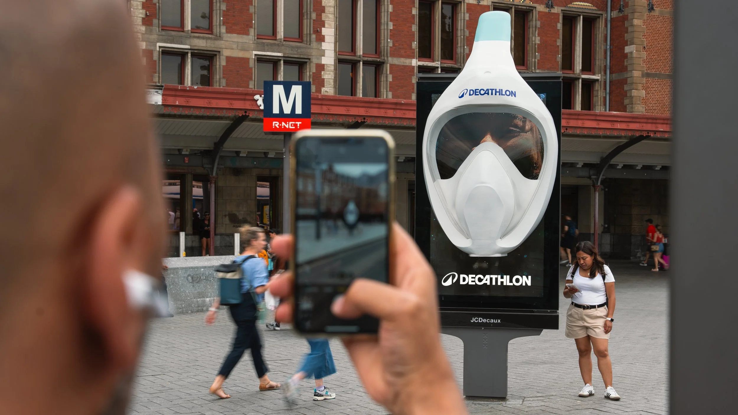 Een man neemt een foto van een bushokje met een advertentie van Decathlon, terwijl binnen een vrouw zich op haar telefoon bevindt en andere mensen door het stadscentrum lopen.