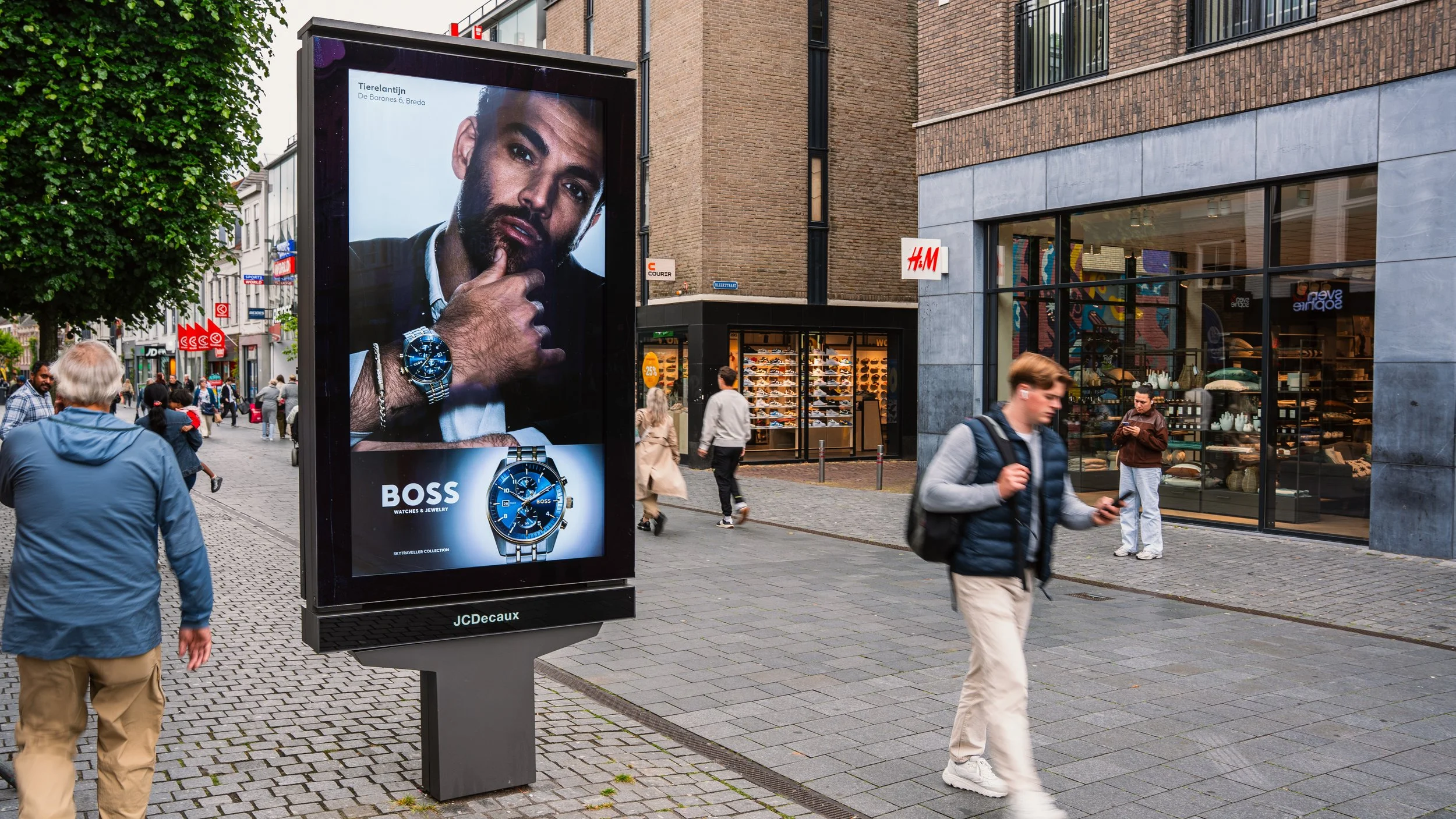 Straatbeeld met mensen wandelend, een digitale reclamezuil die een horloge van het merk Boss adverteert, en winkels zoals H&M op de achtergrond.