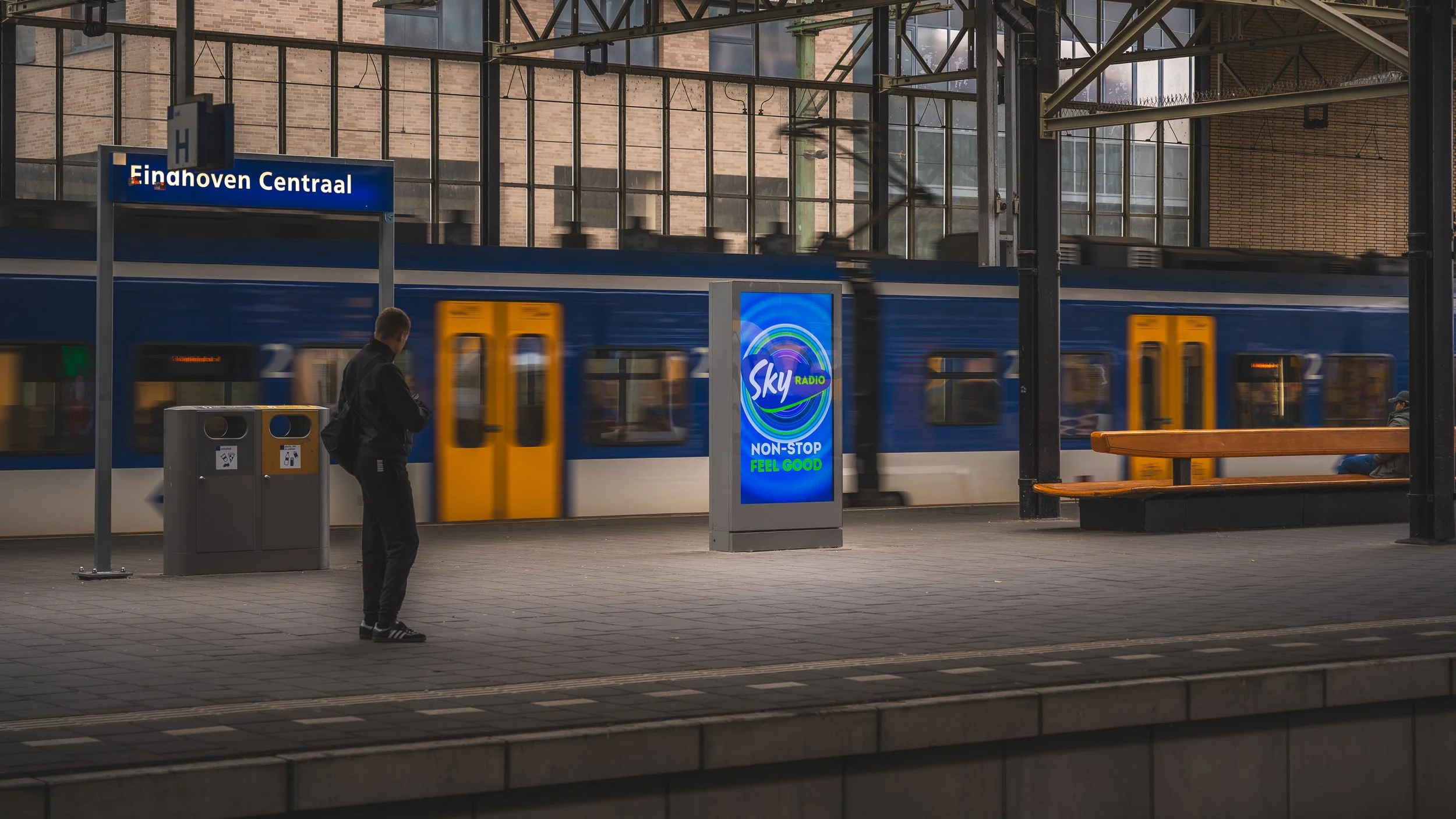Een persoon wacht op een perron bij station Eindhoven Centraal terwijl een blauwe treindienst door het station rijdt, met een digitale reclamezuil en een bankje op de achtergrond.