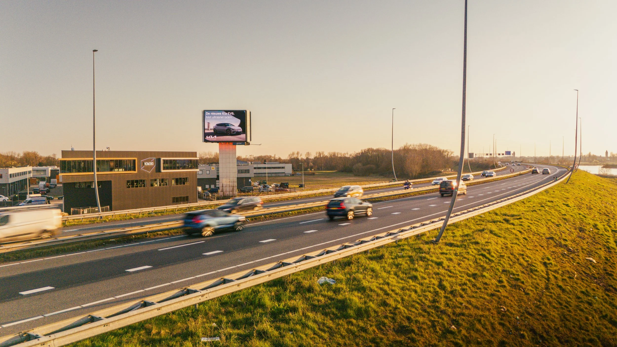 Snelweg met meerdere auto's in beweging, een billboard met een auto en tekst, en een gebouw met een logo en ramen. Het is een zonnige dag.