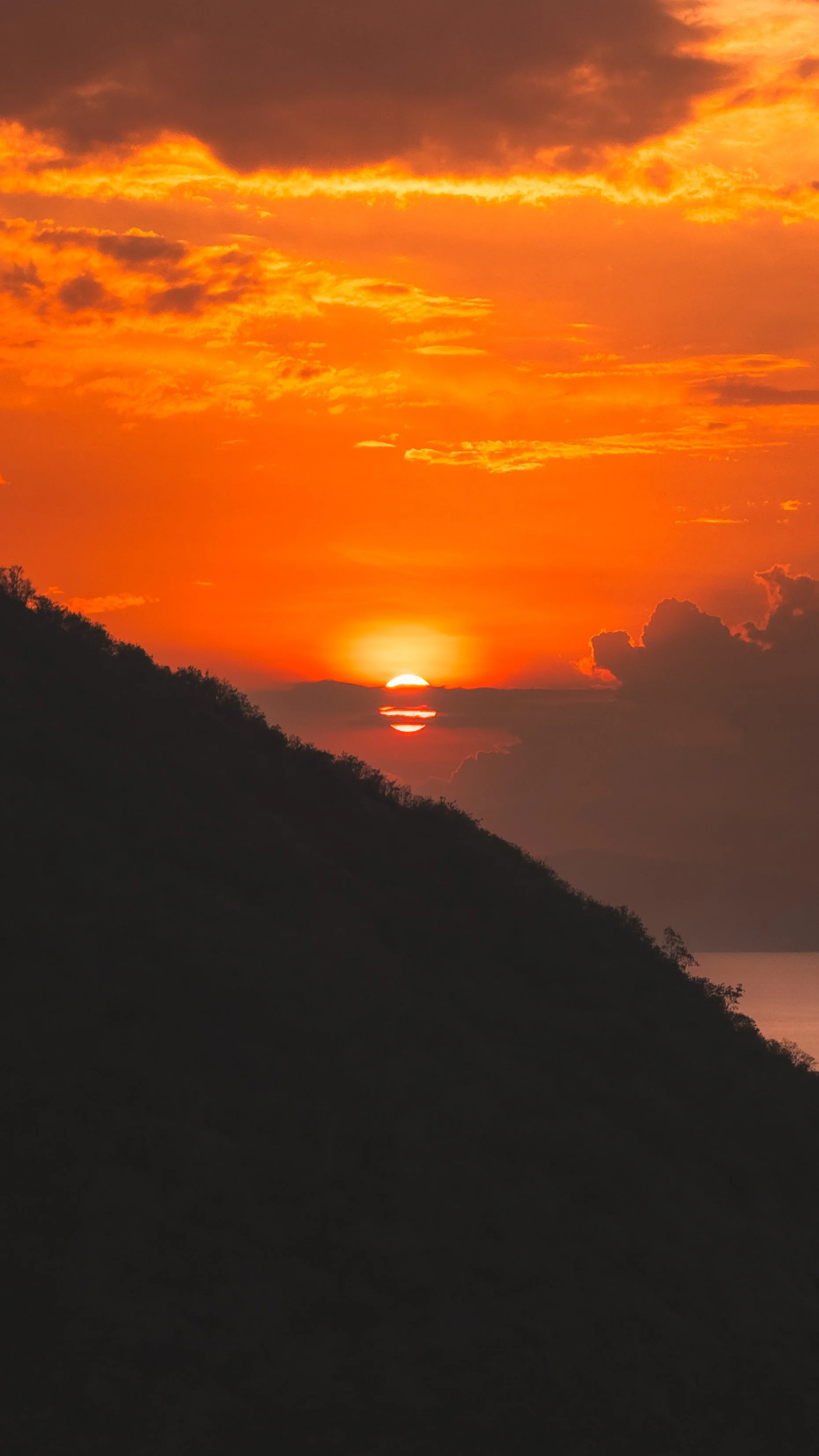 Een kleurrijke zonsondergang boven een heuvel met een bewolkte hemel in oranje en geel tinten.
