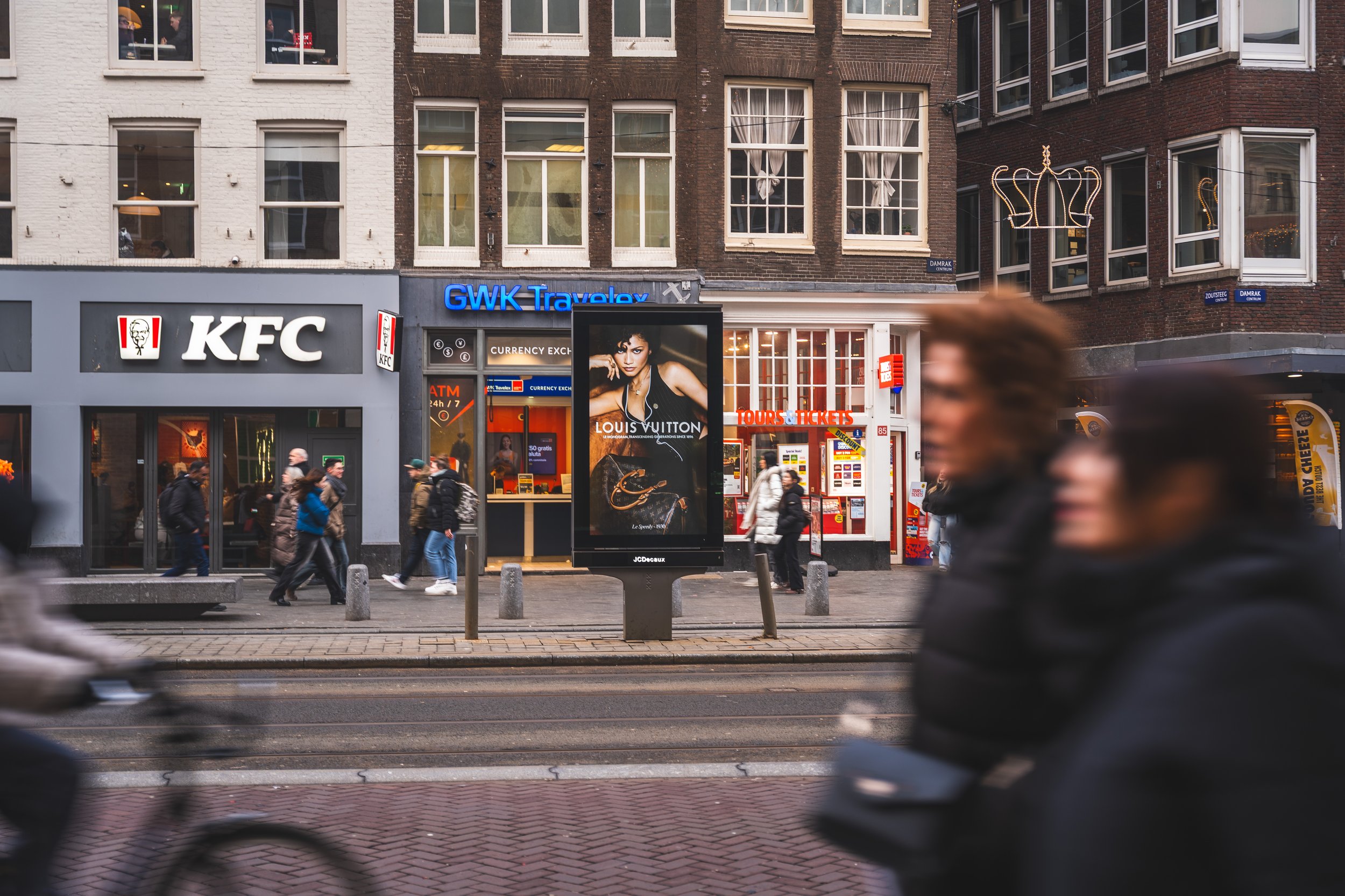 Straatbeeld met winkelpanden en mensen die lopen, inclusief een digital billboard met een reclame voor Louis Vuitton en een KFC-restaurantaandeel achter glas. Bussen en fietsers passeren, in een stedelijke omgeving.
