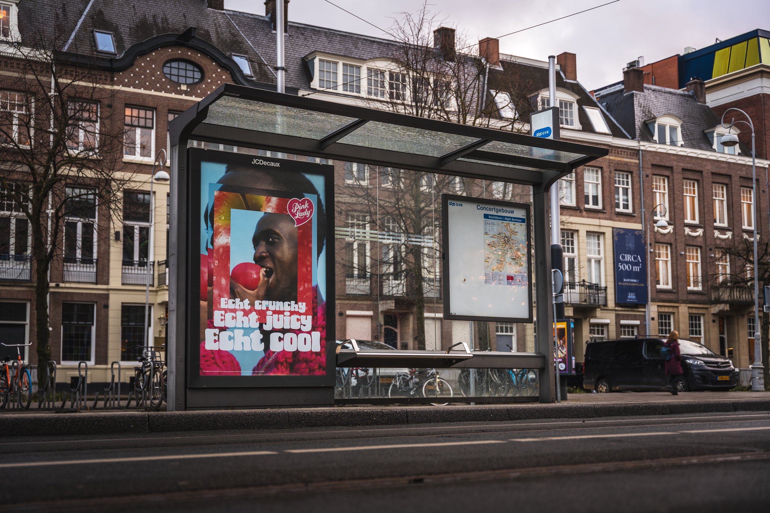 Een bushalte met een advertentie van een vrouw die een rode appel bij haar mond houdt, met teksten in het Nederlands die 'Echt crunchy, Echt juicy, Echt cool' zeggen, en een map van het concertgebouw in Amsterdam op de achterkant.