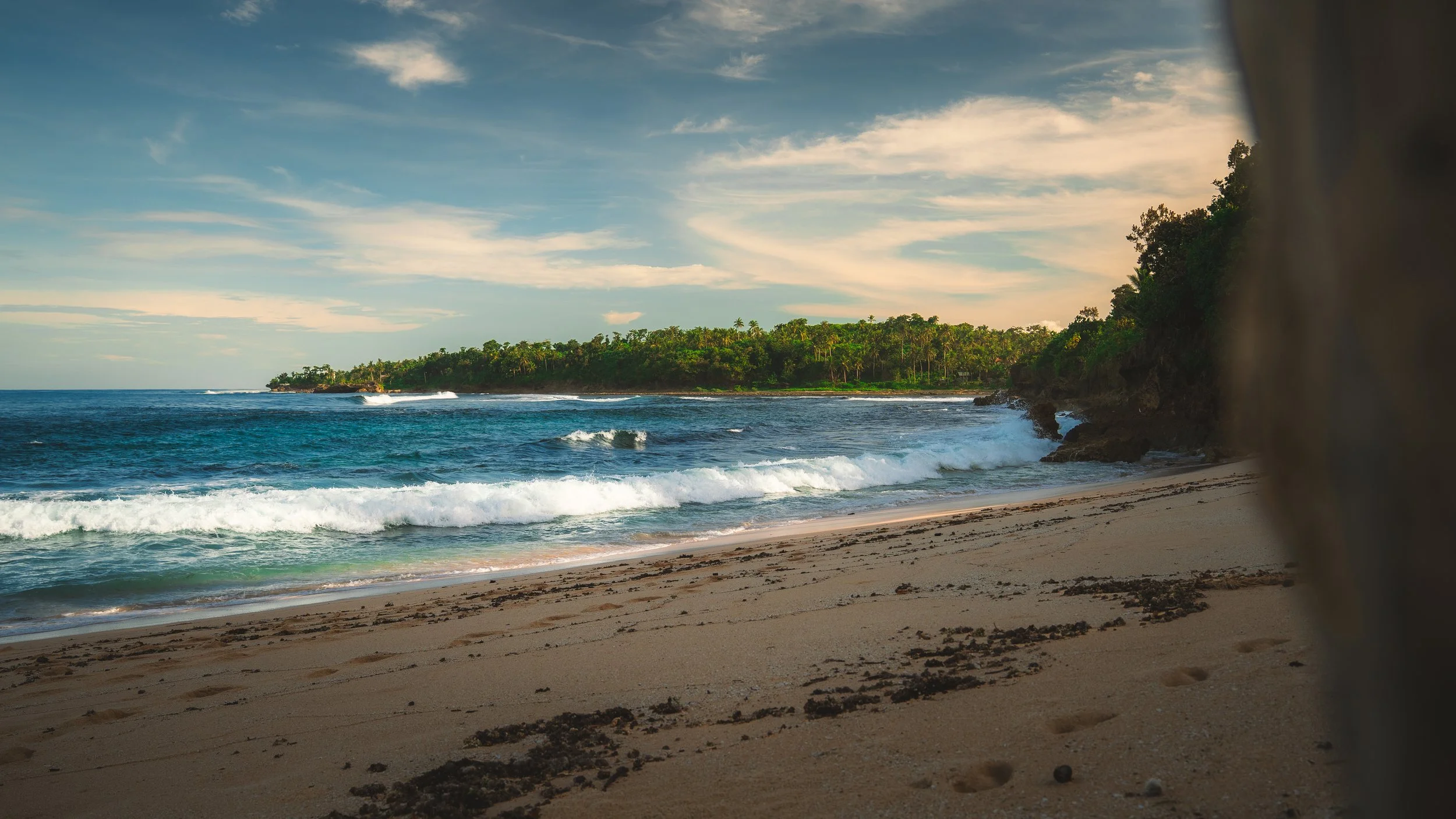 Een zomers strand met zand, branding en een bosrijke kustlijn onder een bewolkte blauwe hemel.