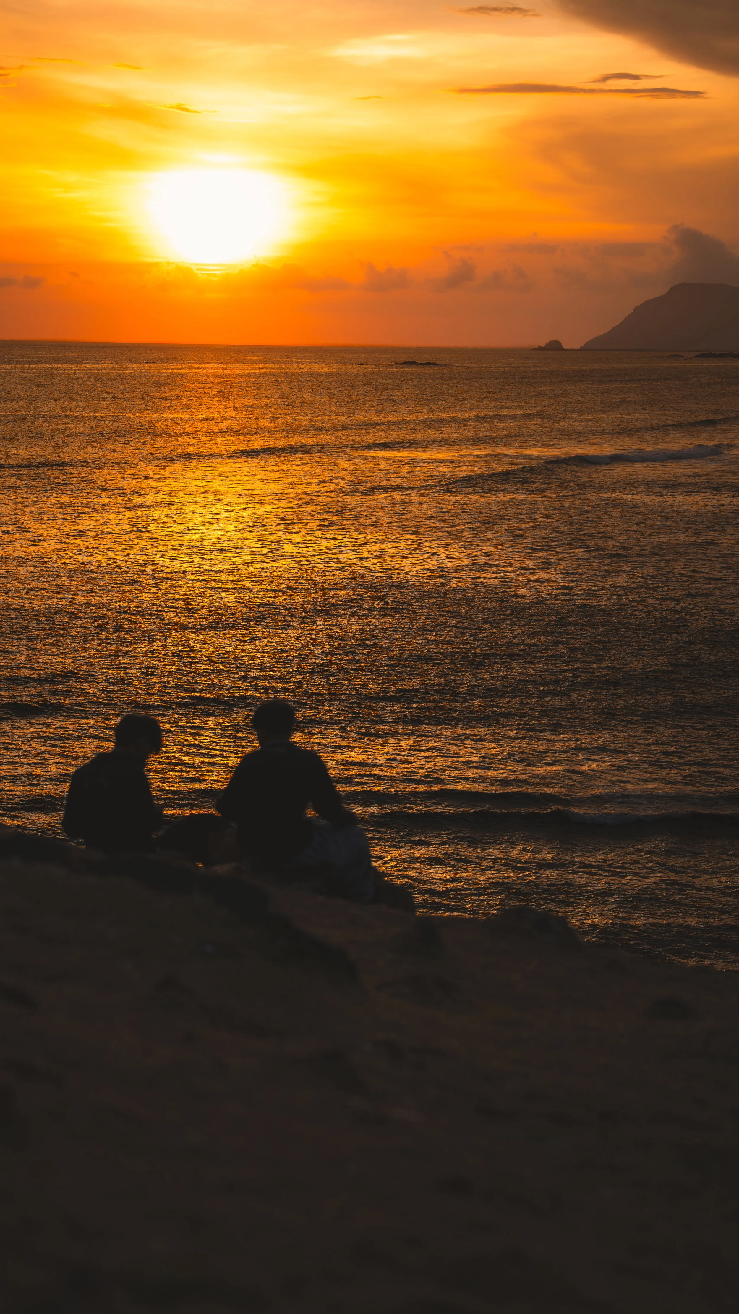 Twee mensen die op een rots bij de zee zitten tijdens een zonsondergang, met de zon die ondergaat en het licht dat reflecteert op het water.