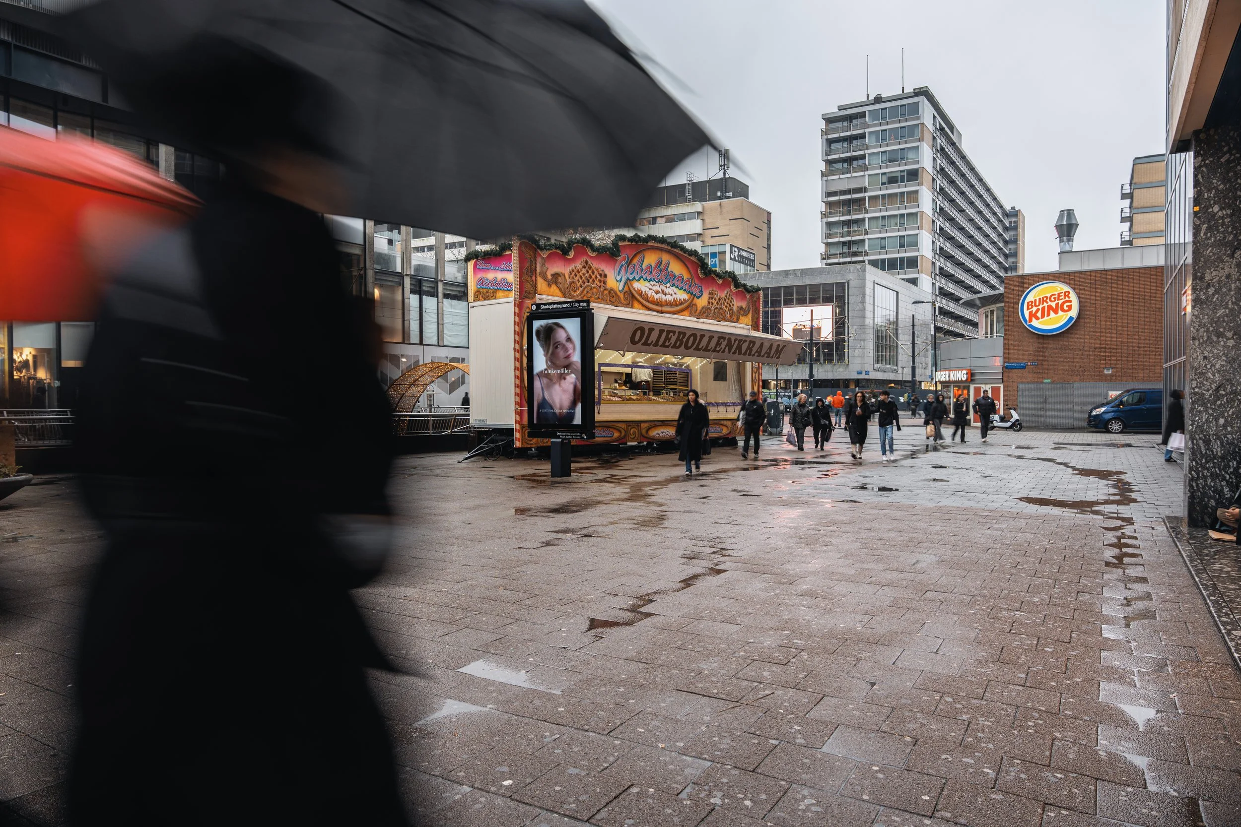 Stadsscene met mensen onder paraplu's op een natte markt, reclamebord voor oliebol en een Burger King restaurant op de achtergrond.