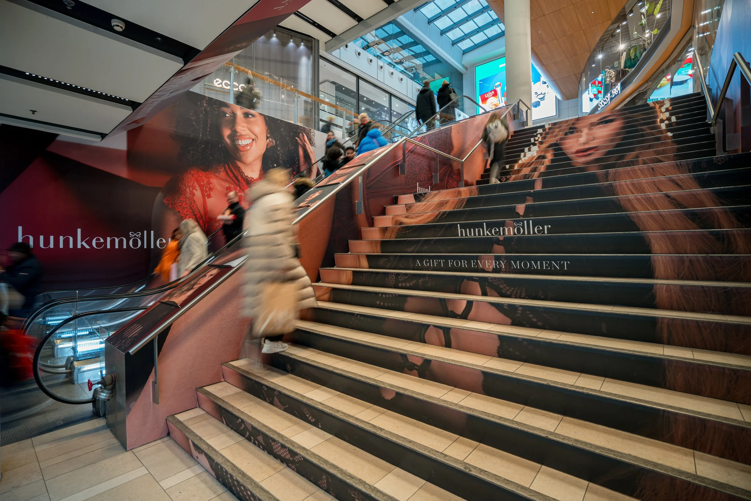 Binnenkant van een winkelcentrum met een trap en reclame voor Hunkemöller, met grote foto's van vrouwen en mensen die de trap op en af lopen.
