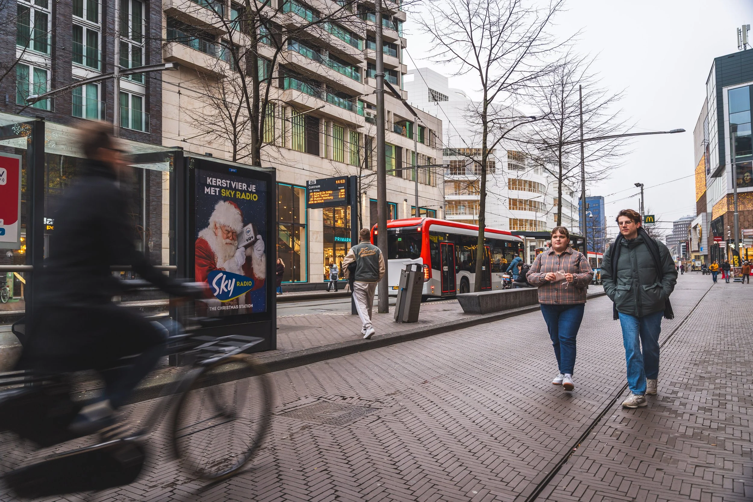 Stadsplein met mensen, een fiets, een bus, een bushokje met kerstadvertentie, gebouwen en bomen in de winter, met een bewolkte hemel.