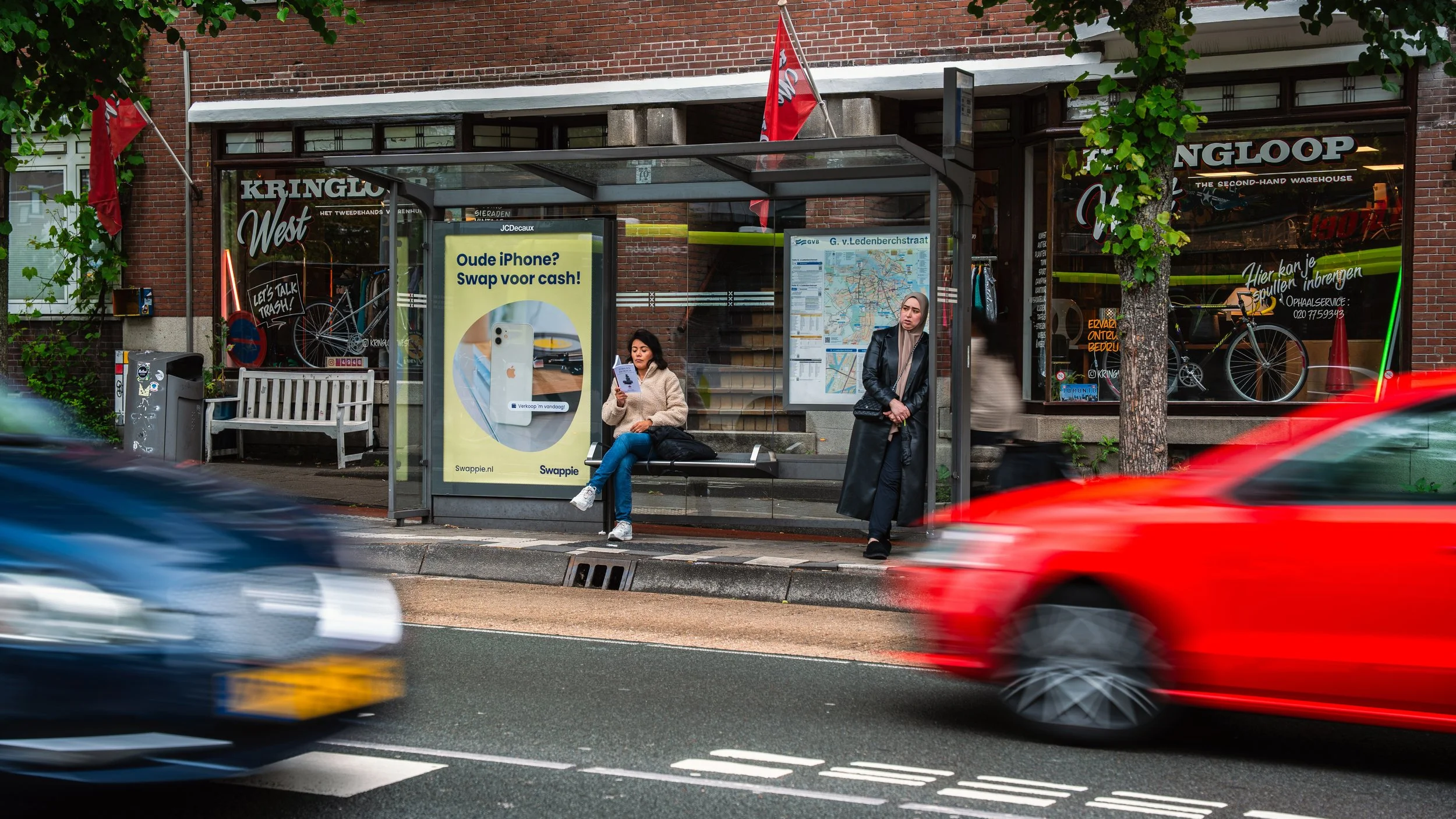 Twee vrouwen wachten bij een bushalte in een stadswijk. Eén vrouw zit op de bank, de andere staat ernaast. In de achtergrond is een winkel met fietsen en reclameborden te zien, met auto's die door de straat rijden.