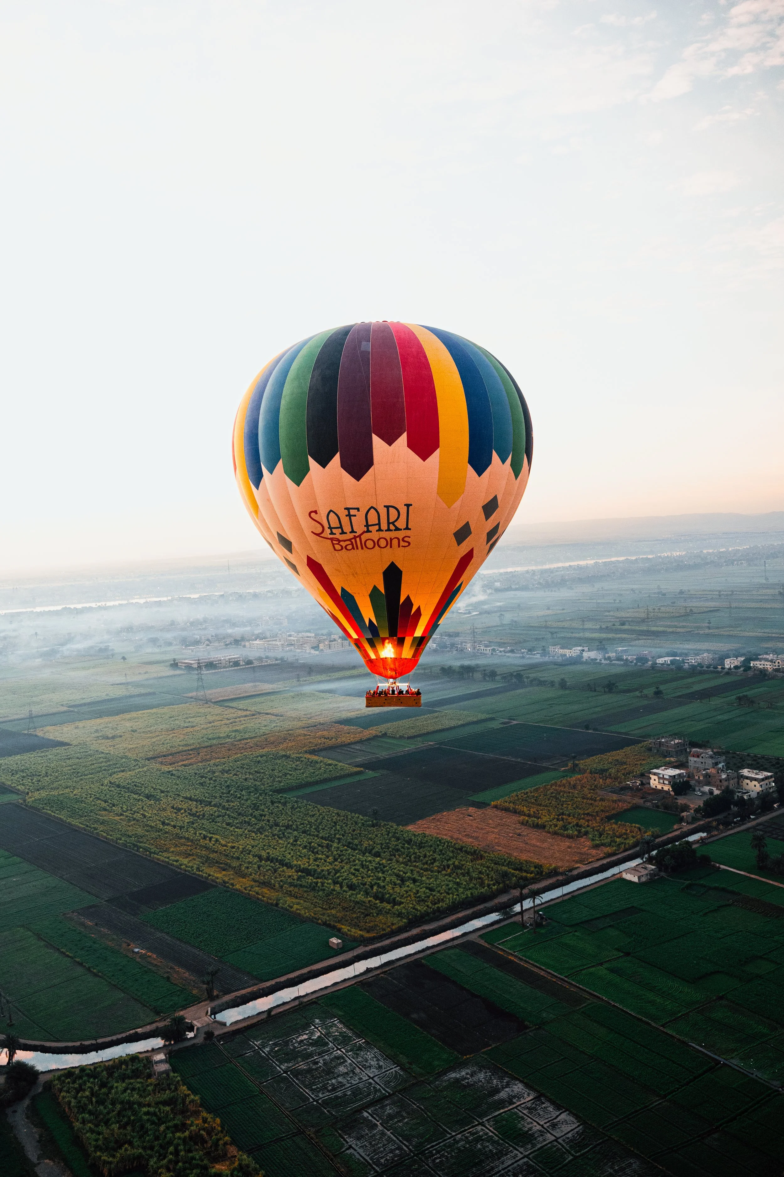 Een kleurrijke luchtballon met de tekst 'Safari Balloons' zweeft boven een vlak landschap met velden en huizen.
