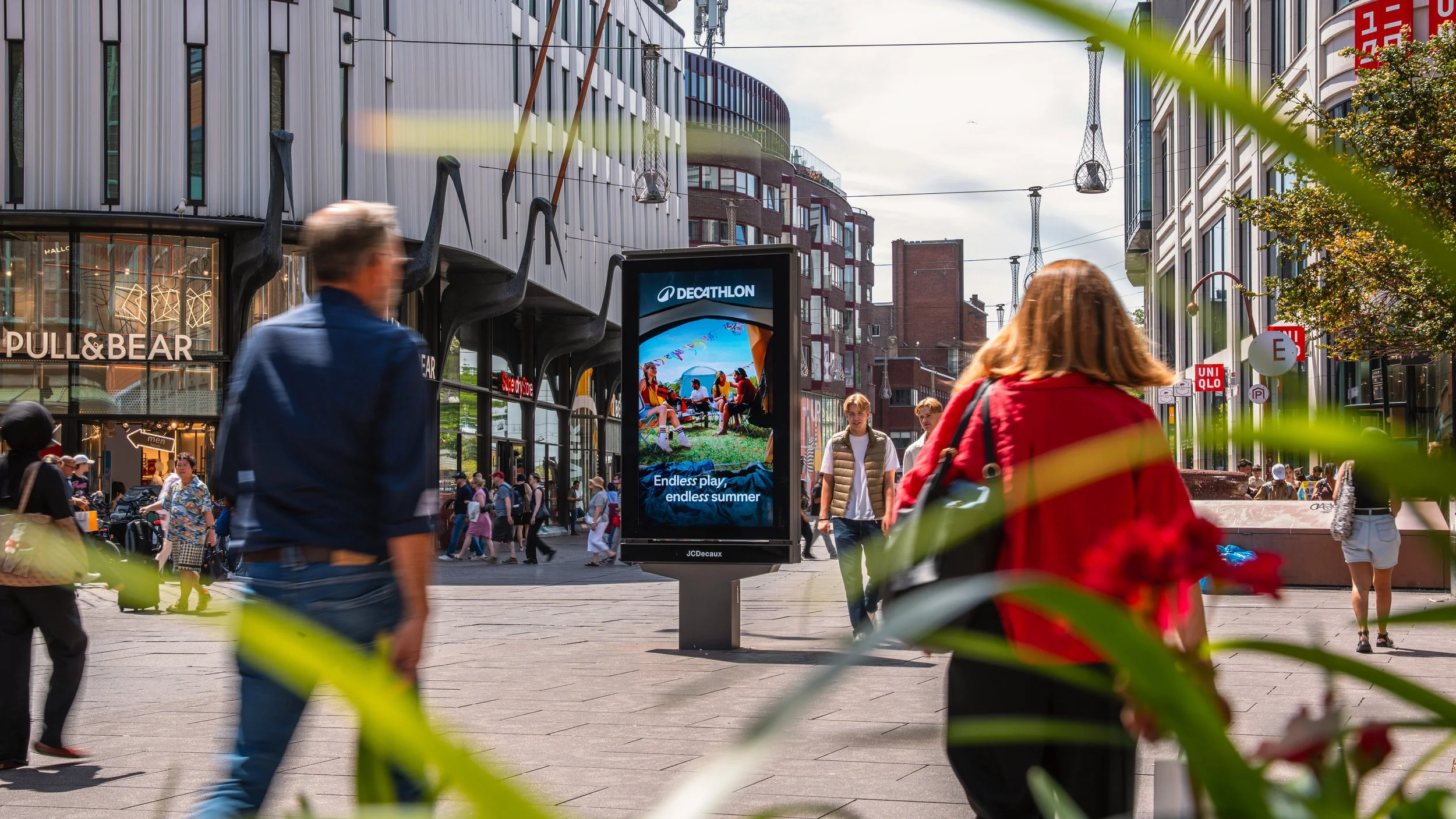 Een drukke stadsstraat met mensen die lopen, moderne gebouwen en een digitale reclamezuil die advertentietekst toont.