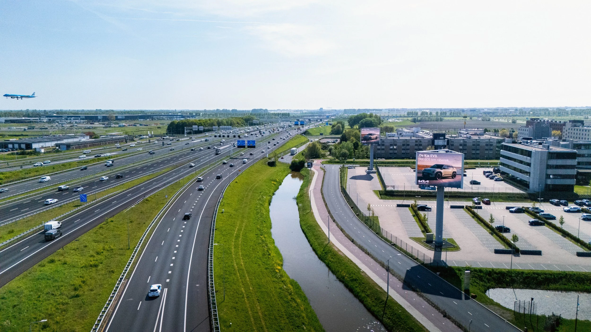 Luchtfoto van een snelweg met meerdere rijstroken vol verkeer, een rivier die parallel aan de snelweg loopt, en een bedrijfscomplex met billboards en parkeergelegenheid. In de lucht vliegt een vliegtuig.