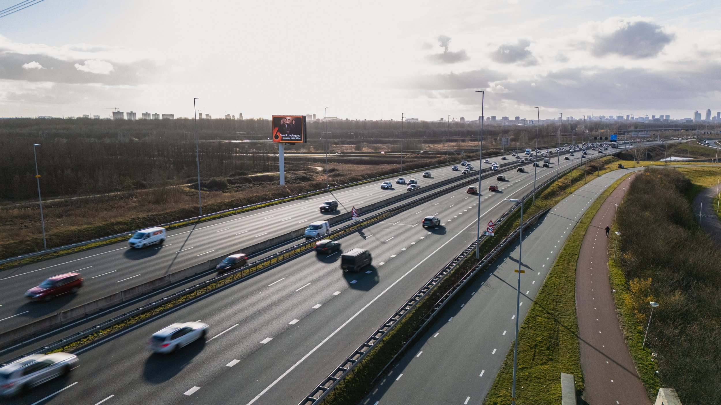 Autos rijden op een snelweg met meerdere rijstroken en een weg ernaast met wandel- en fietspaden, onder een bewolkte hemel.