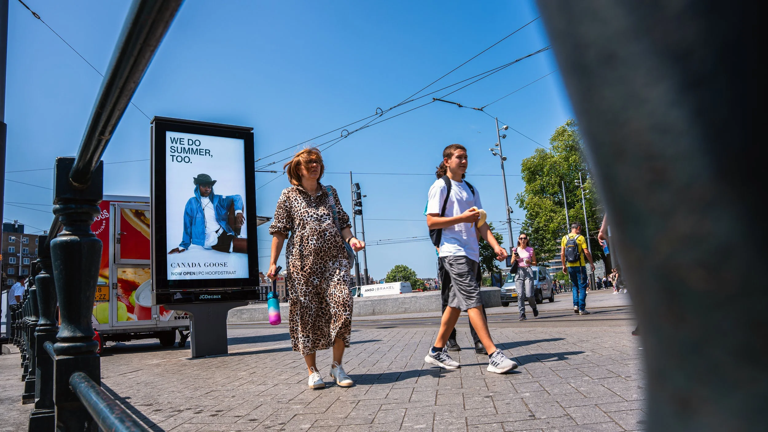 Mensen lopen over een stedelijk plein op een zonnige dag, met een digitale affiches aan de achtergrond en een blauwe lucht.