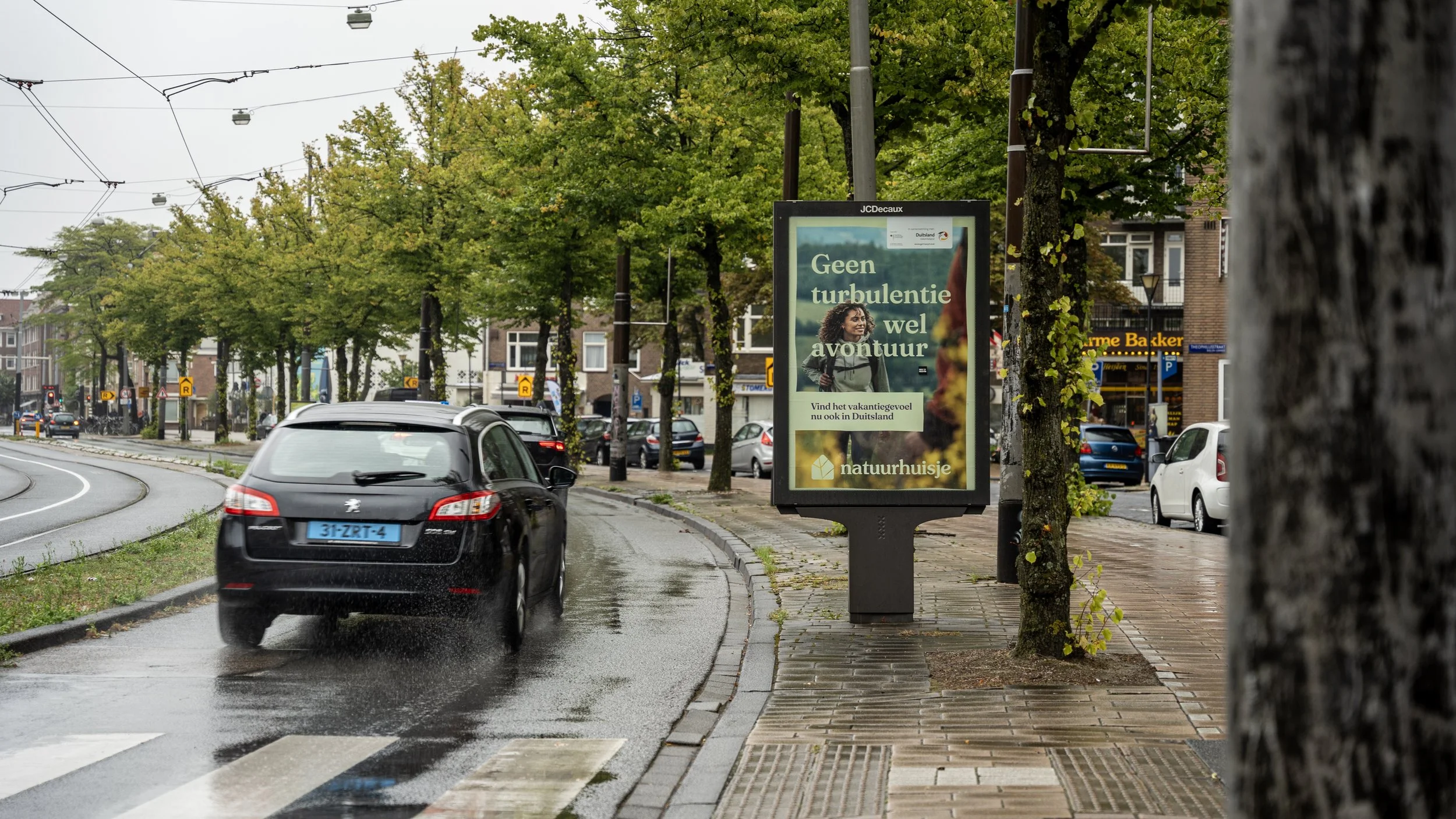 Een regenachtige straat in Nederland met een auto op de rijbaan, een bushaltestructuur met een reclamebord, bomen met groene bladeren, en gebouwen op de achtergrond.