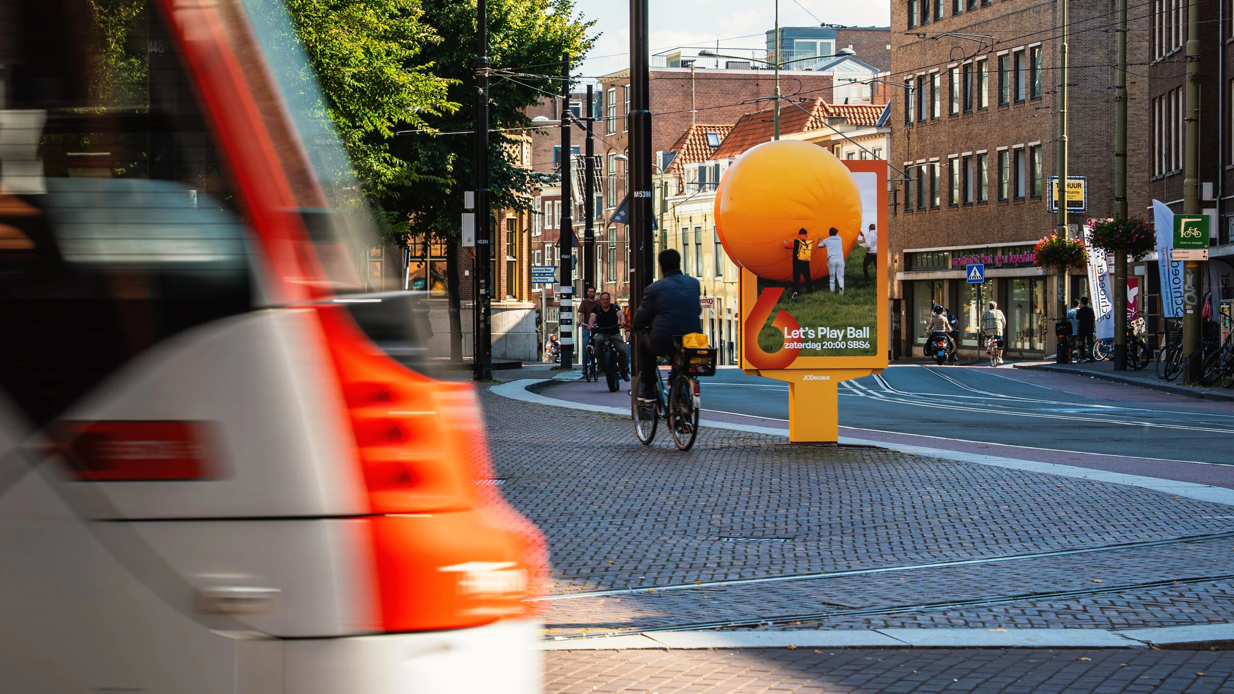 Een stadsscene met fietsers, een opvallend oranje advertentiebord met een grote bal en tekst, en een rood-witte tram die door de straat rijdt. Mensen lopen en fietsen op het trottoir, met gebouwen op de achtergrond.