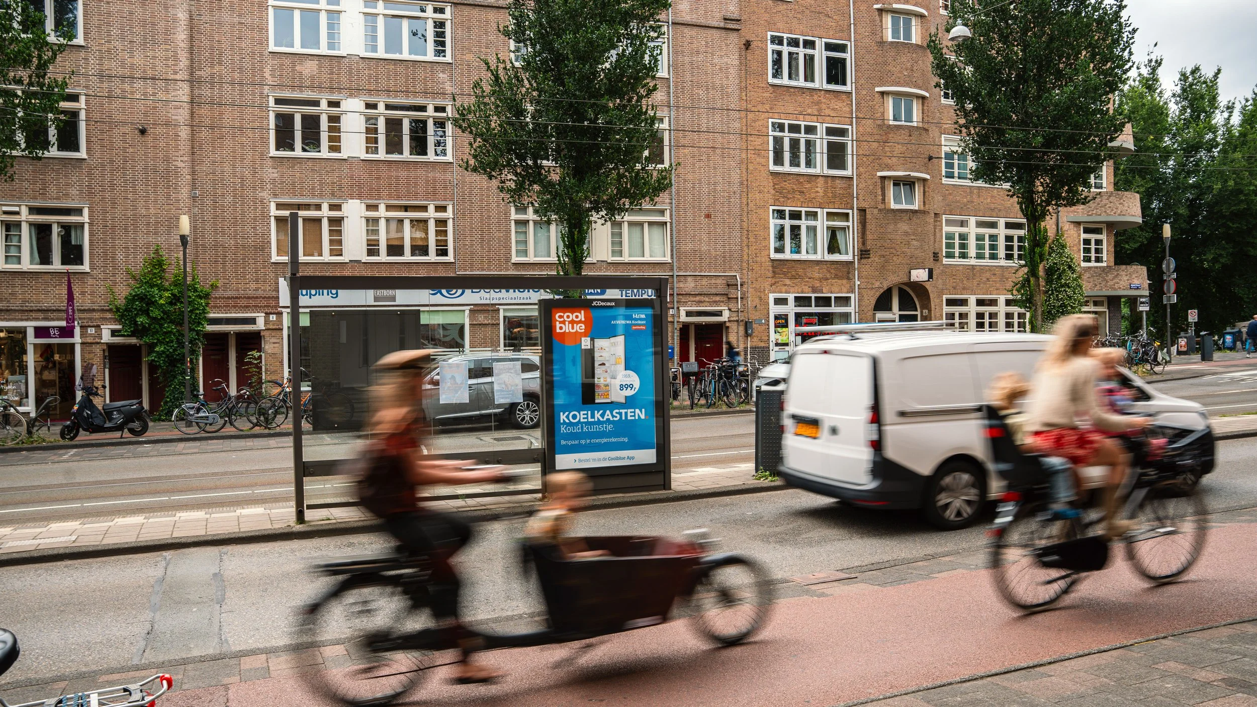 Winkelcentrum of kantoorgebouw met fietsenstalling voor de ingang en reclameborden. Mensen fietsen langs een bushokje op een stadsstraat met tramlijn.