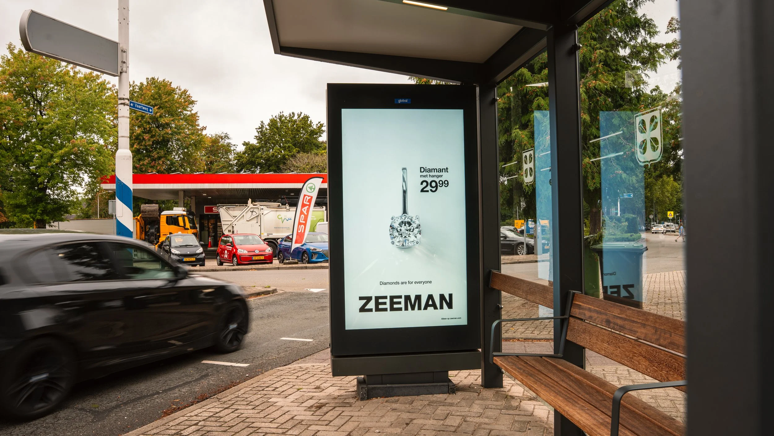 Bushokje met digitale advertentie voor een diamant met hanger, prijs 29,99 euro, door Zeeman. Op de achtergrond zijn bomen, een benzinestation en geparkeerde auto's zichtbaar,