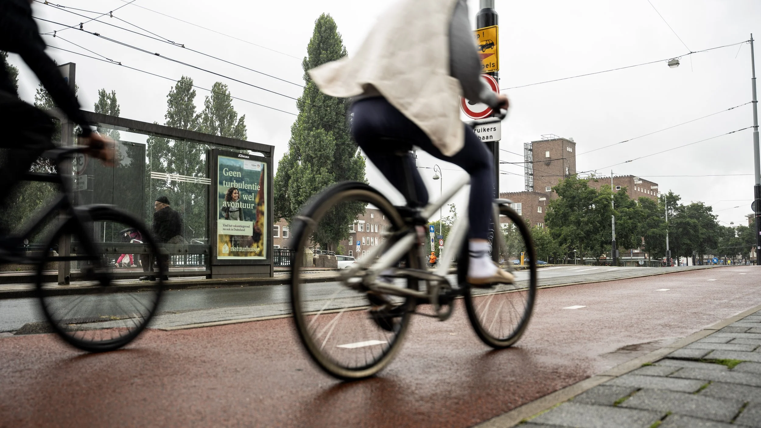 Een persoon fietst op een rode fietsstrook op een straat, in de achtergrond zijn bomen en gebouwen te zien en een bushokje met een poster.