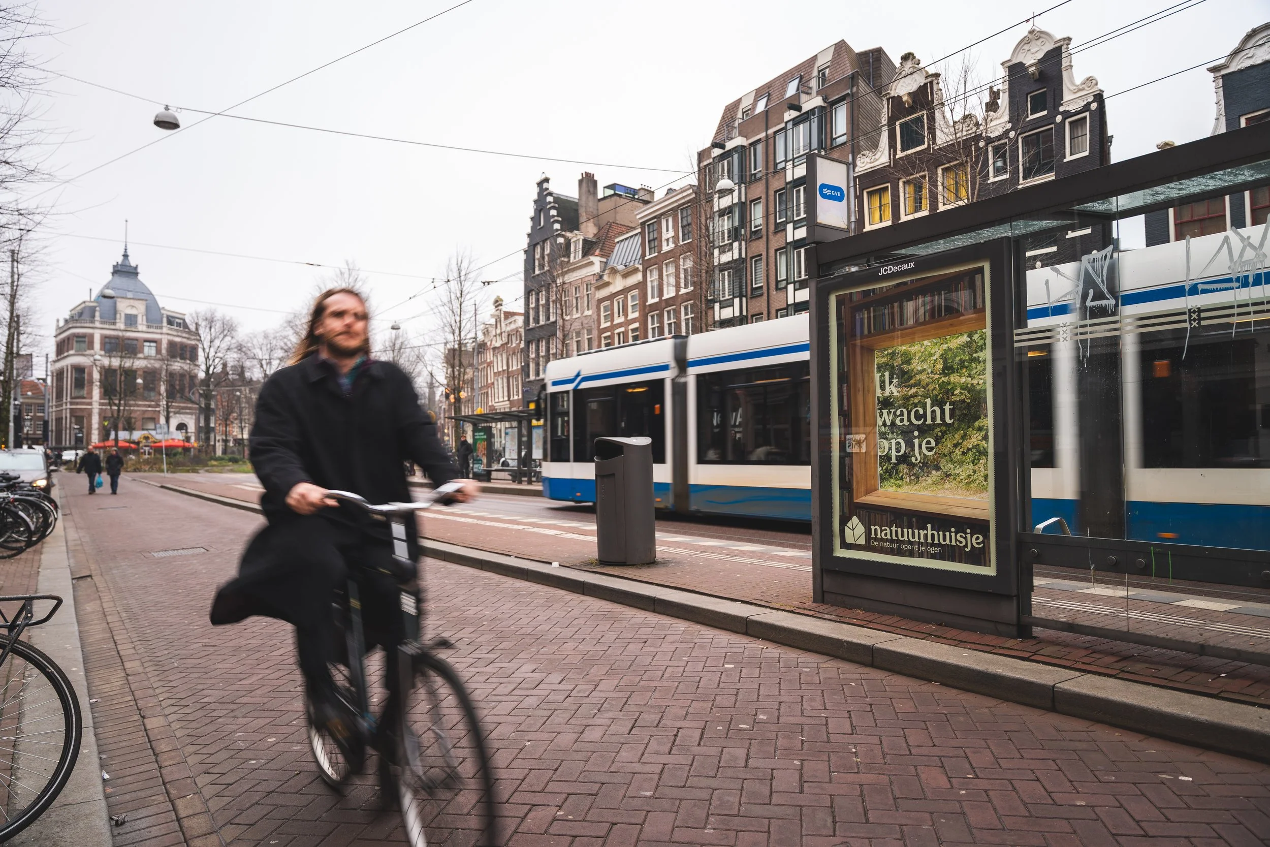Man fietst langs tramhalte met advertentie, tram rijdt door straat met historische gebouwen, bomen zonder bladeren, op bewolkte dag in Amsterdam.