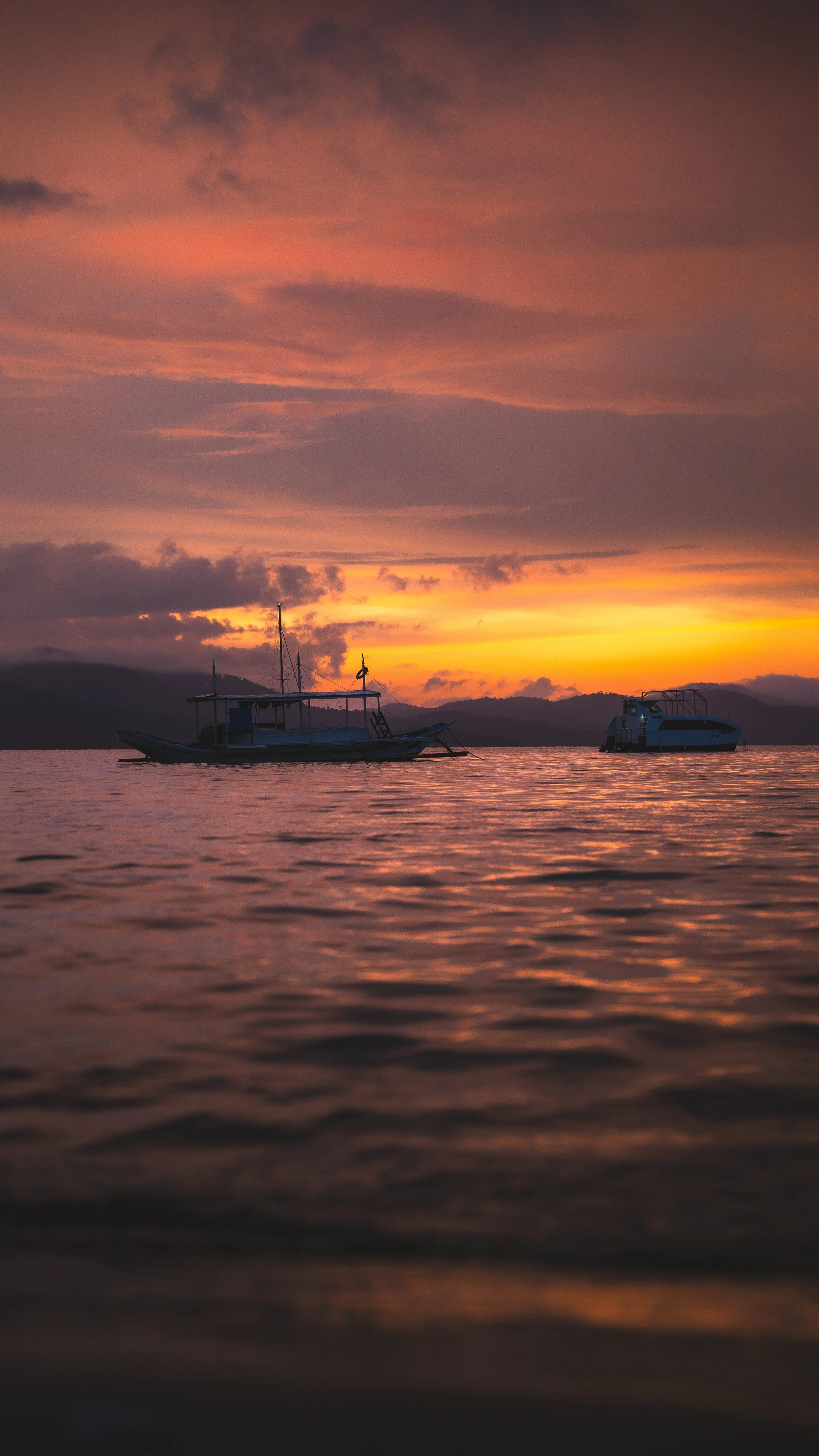 Een foto van een zee bij zonsondergang met twee boten op het water, de hemel is roze en oranje met wolken.