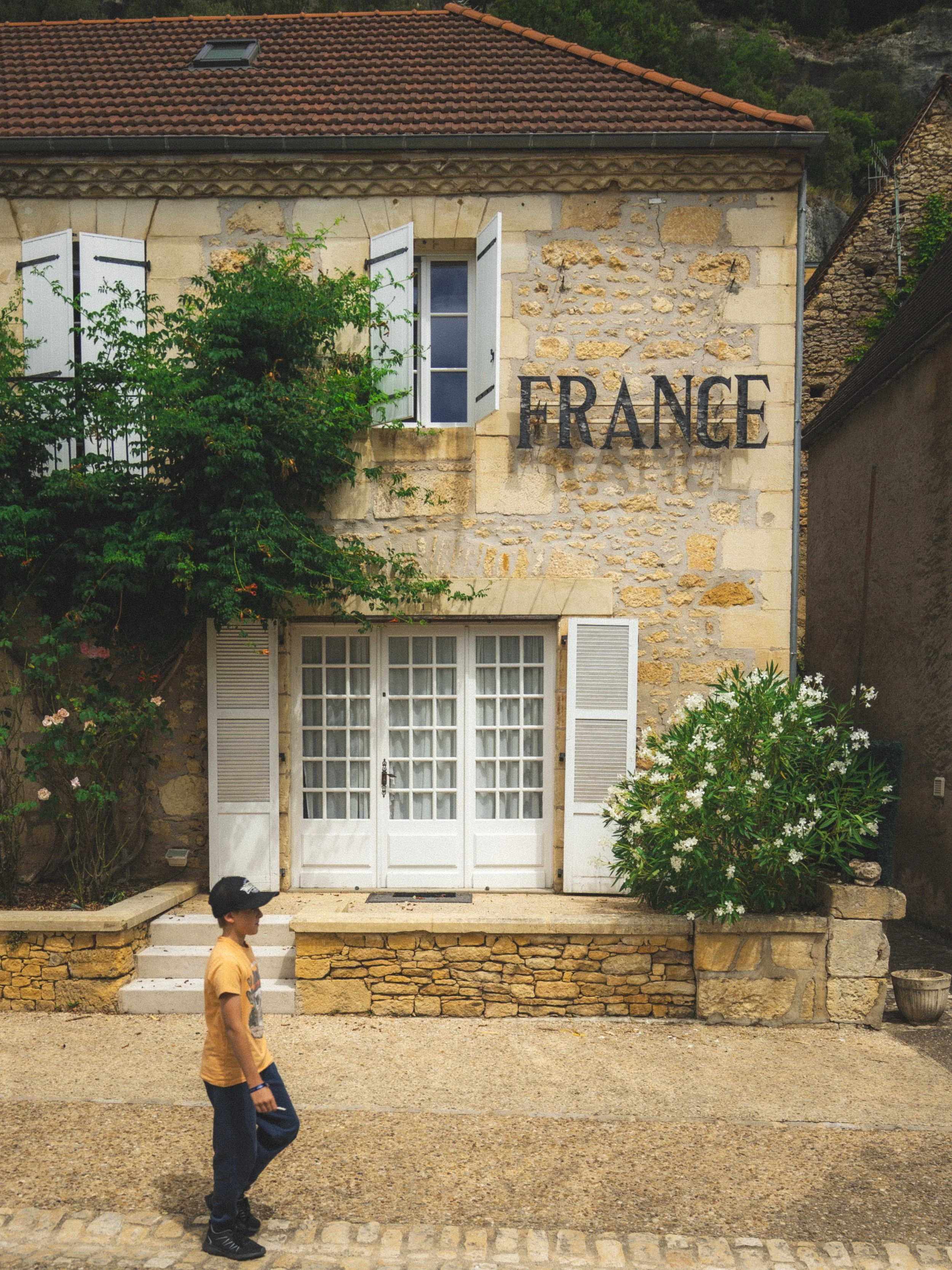 Een oude stenen huis met witte luiken, een grote groene plant aan de gevel en het woord 'FRANCE' erop. Er is een kind dat voorbij loopt, gekleed in een oranje shirt en een zwarte pet.