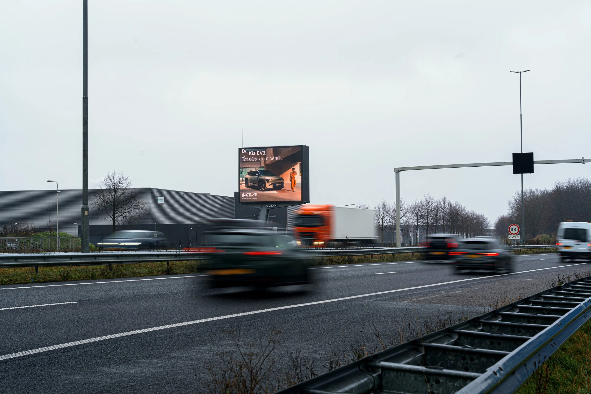Autos rijden snel op een autosnelweg met een reclamebord op de achtergrond, onder een grijze bewolkte hemel.