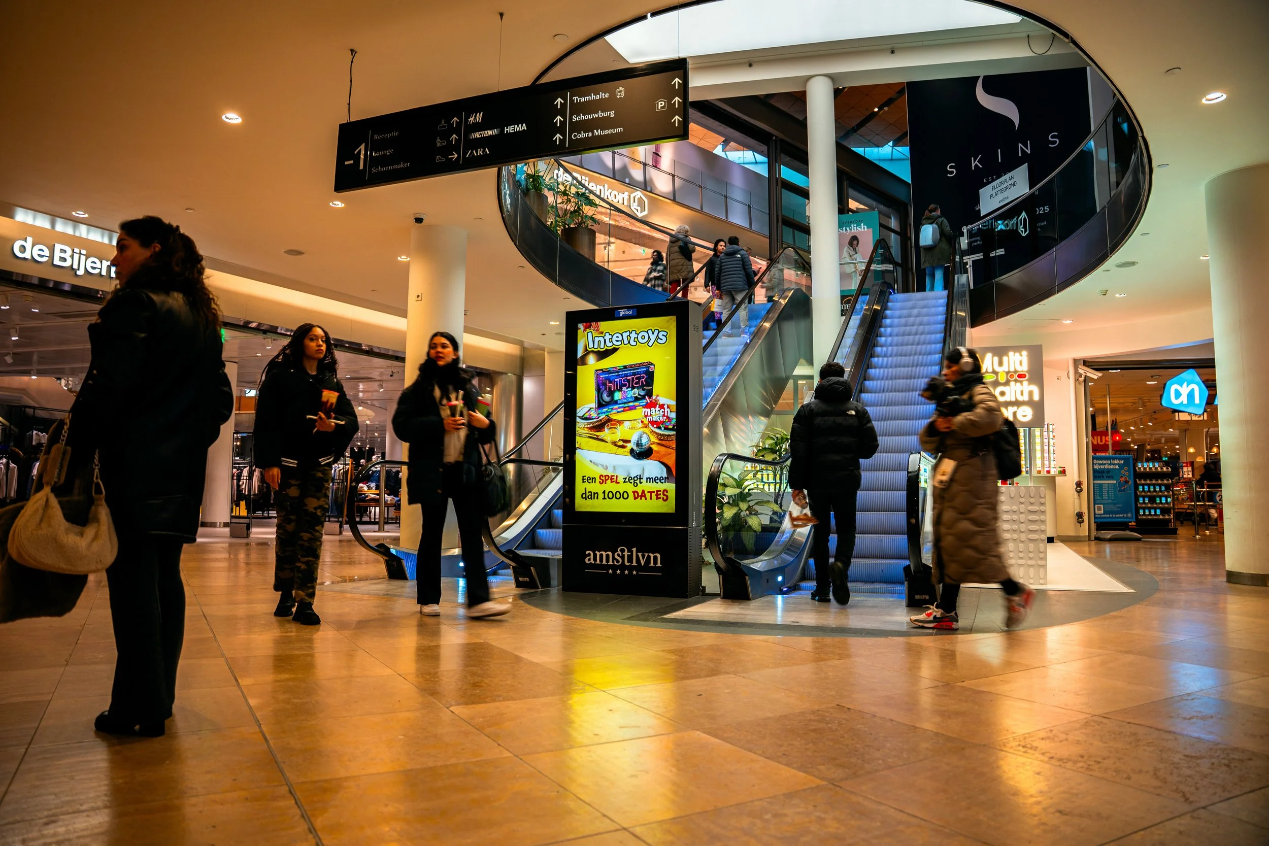 Binnen in een winkelcentrum, met mensen die wachten bij een roltrap en ensceneringsborden met advertenties.