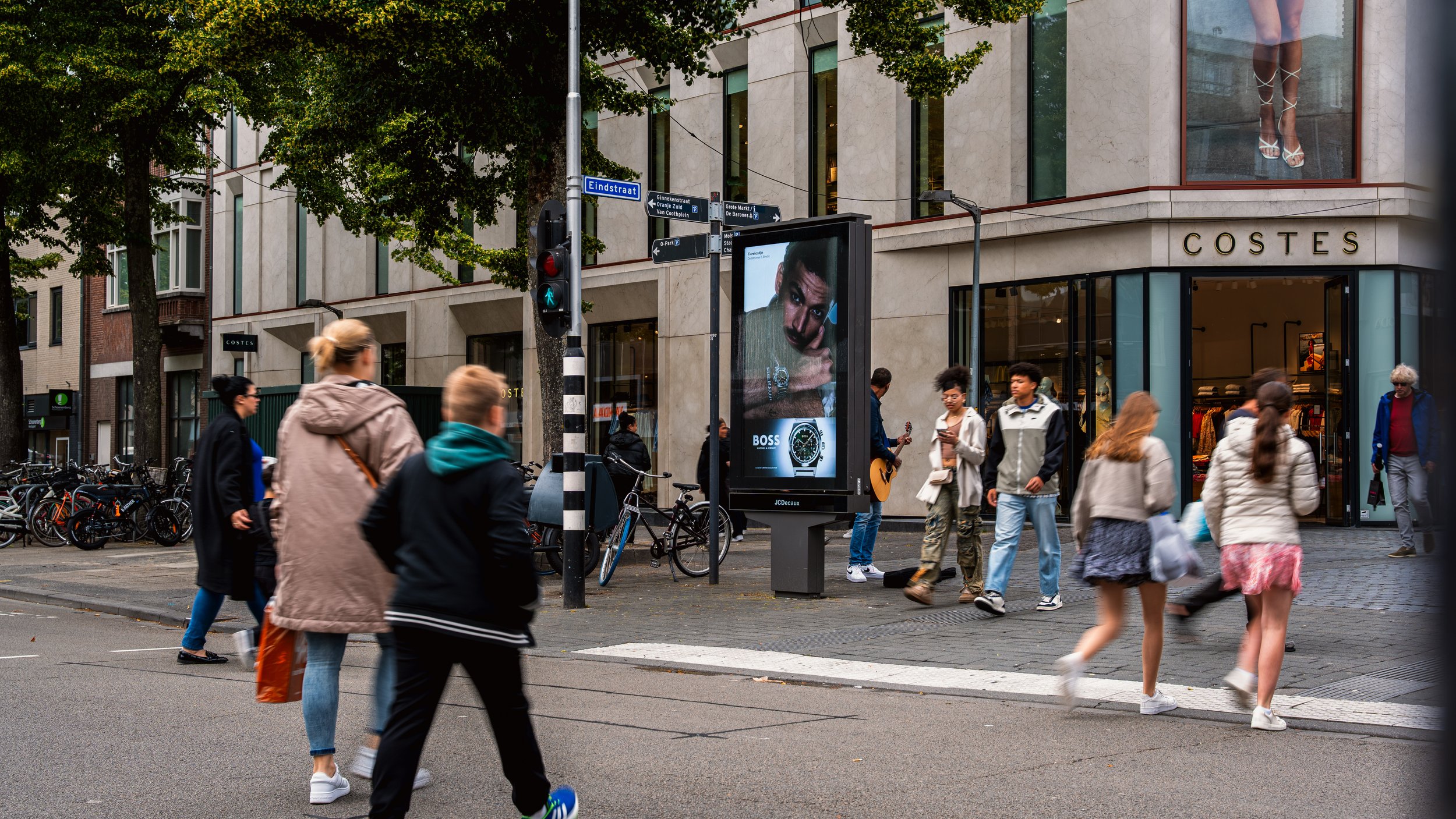 Straatbeeld met voetgangers die de zebrapad oversteken. In het achtergrond is een winkel met de naam 'COSTES' te zien en een digitale billboard met een model en een horloge.