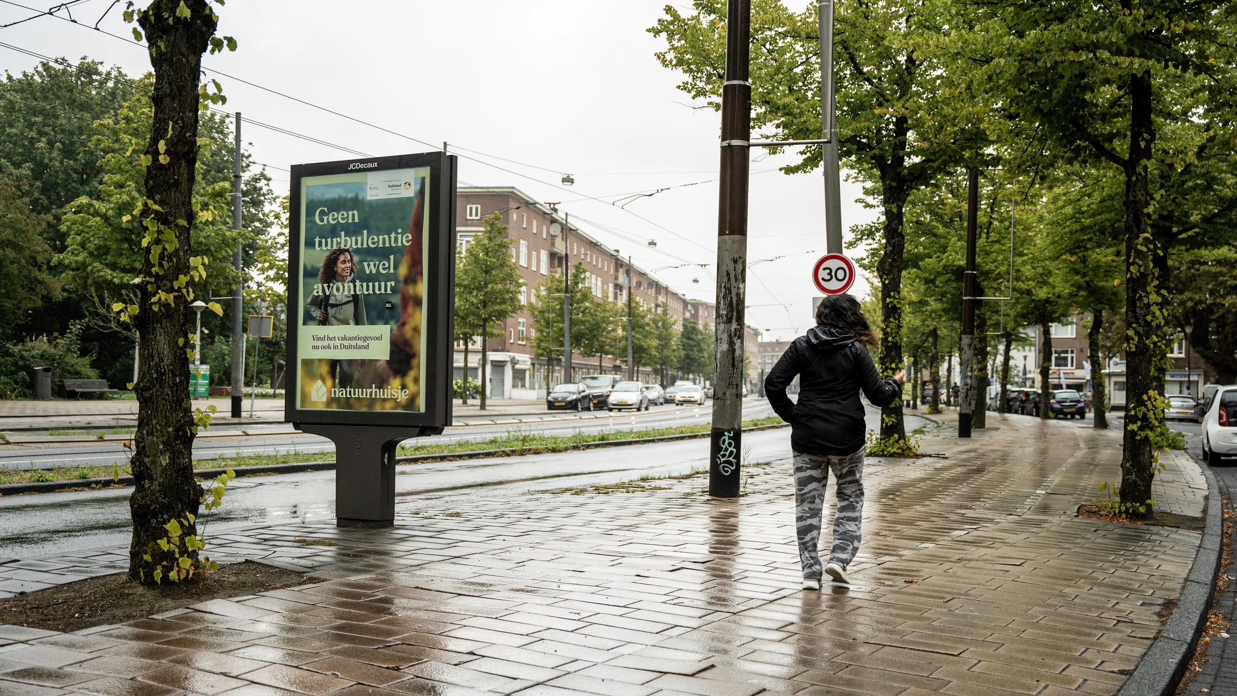 Een vrouw wandelt op een natte, betegelde stoep langs een straat met geparkeerde en rijdende auto's, bomen en een bushokje met een digitale advertentiebord.