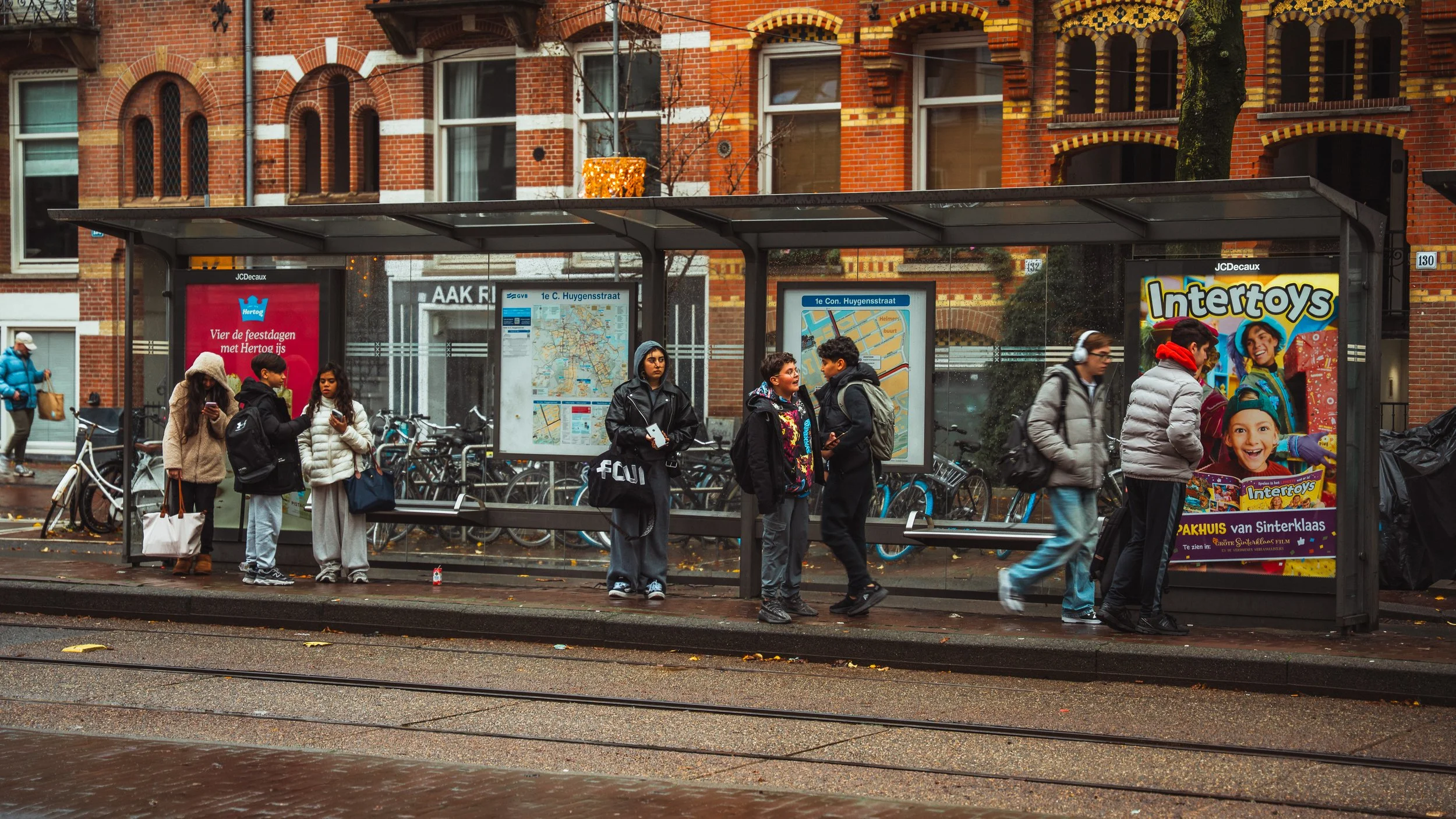 Mensen wachten bij een bushalte op een regenachtige dag, enkele mensen kijken op hun telefoons en anderen lijken te praten.