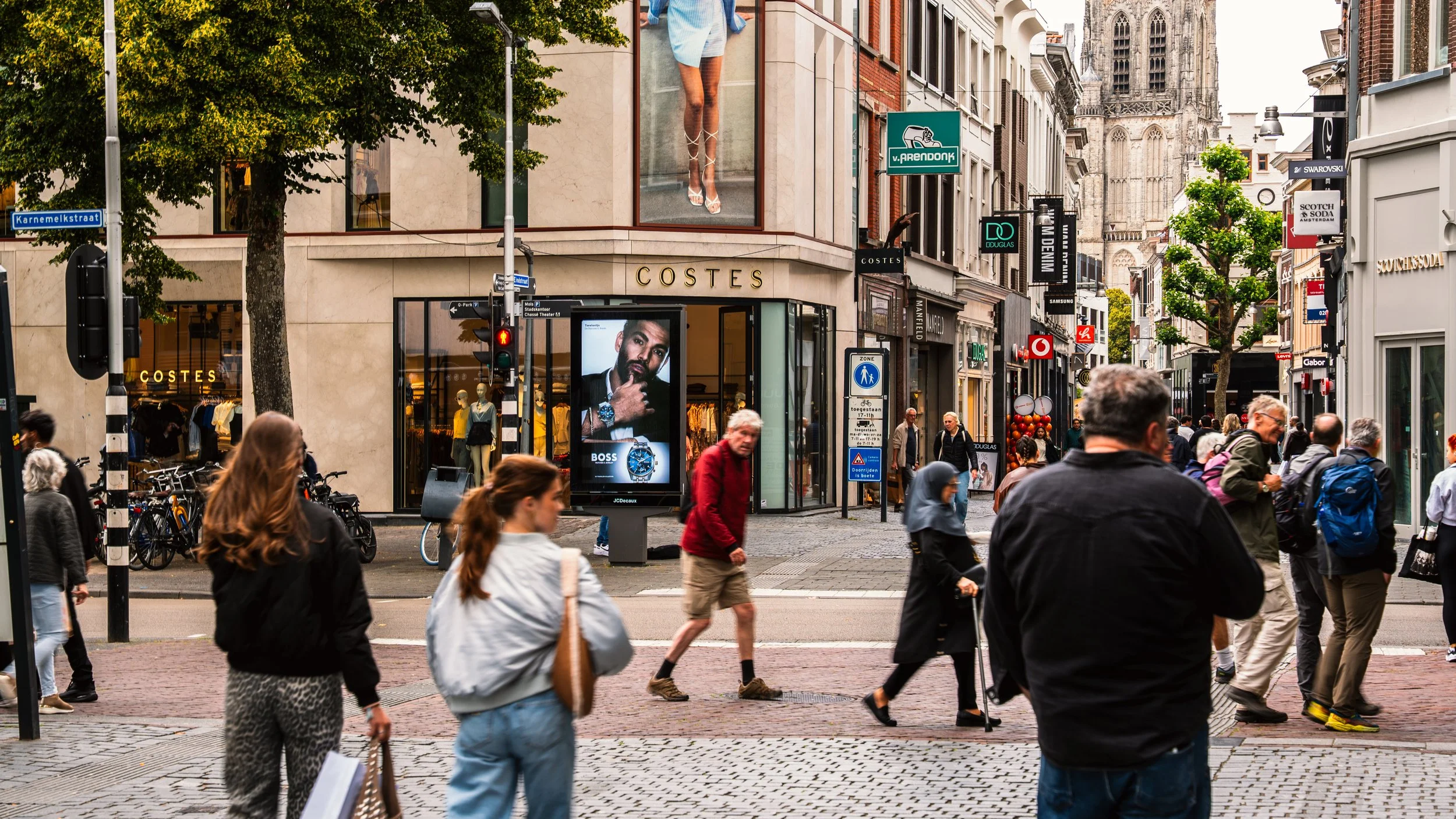 Stedelijke straat met mensen die oversteken, winkels, reclameborden en een kerk op de achtergrond.