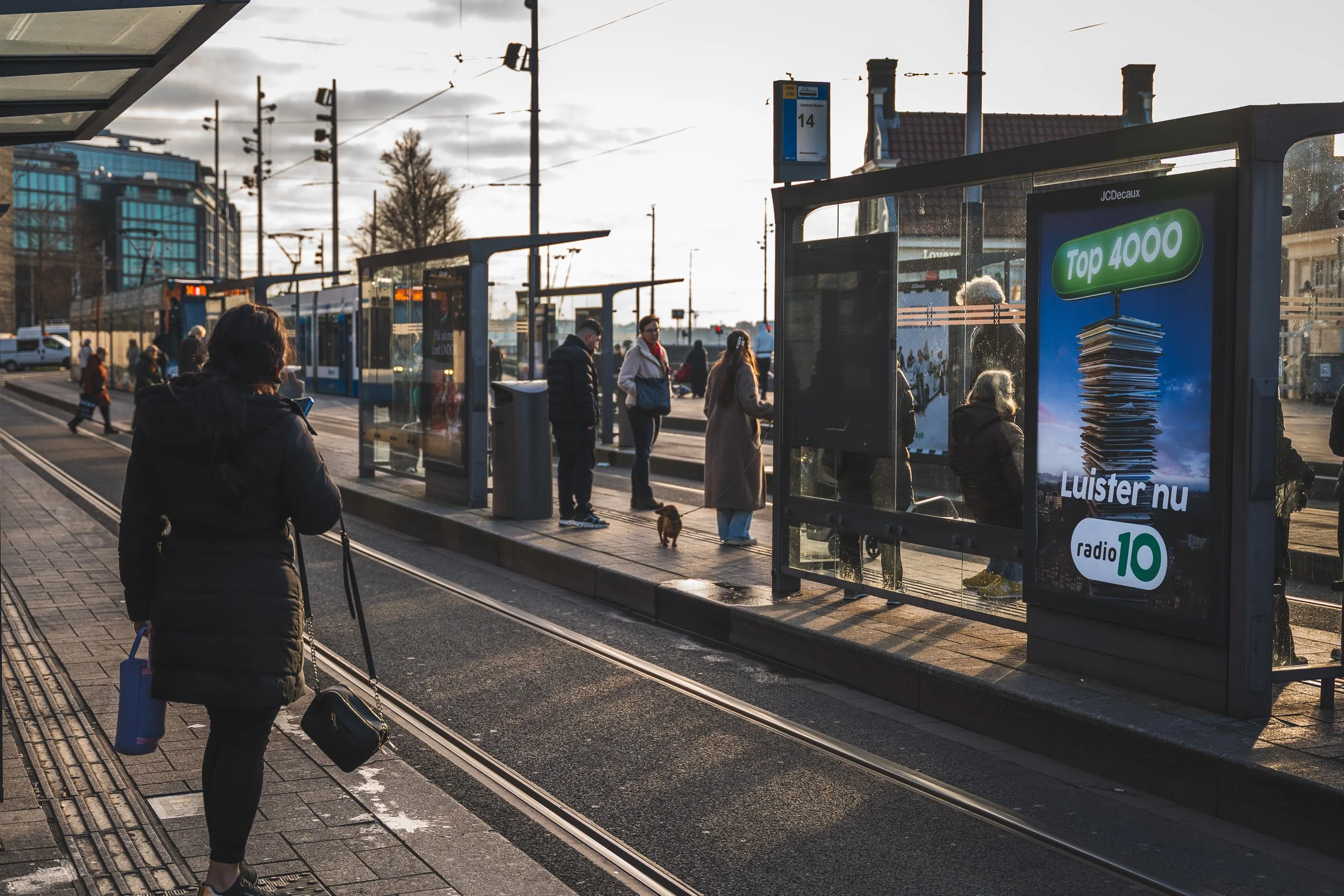 Mensen wachten op een tram bij een bushalte in de ochtend, met een vrouw die een tas en een drinkfles vasthoudt en een hond die naast haar staat. De zon schijnt en de omgeving bevat moderne gebouwen en een overzichtelijke infrastructuur.