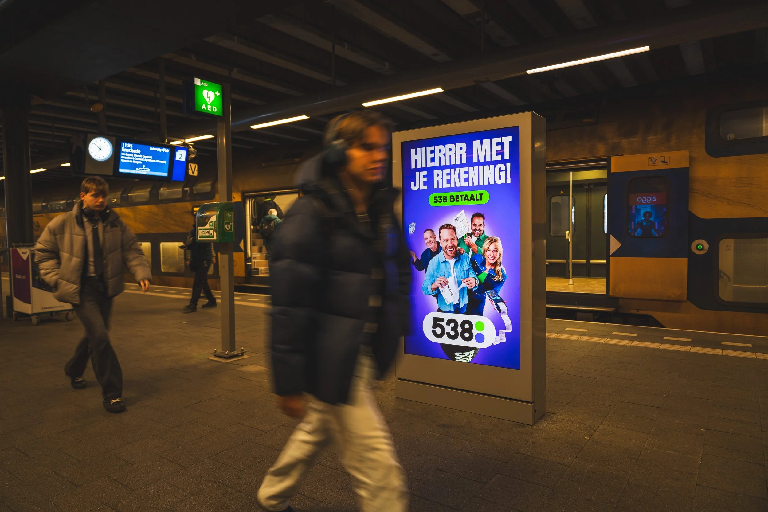 Mensen die over een stationsplatform lopen met een digitale advertentiebord dat een betaalde rekening promoot, in een treinhalte in Nederland.