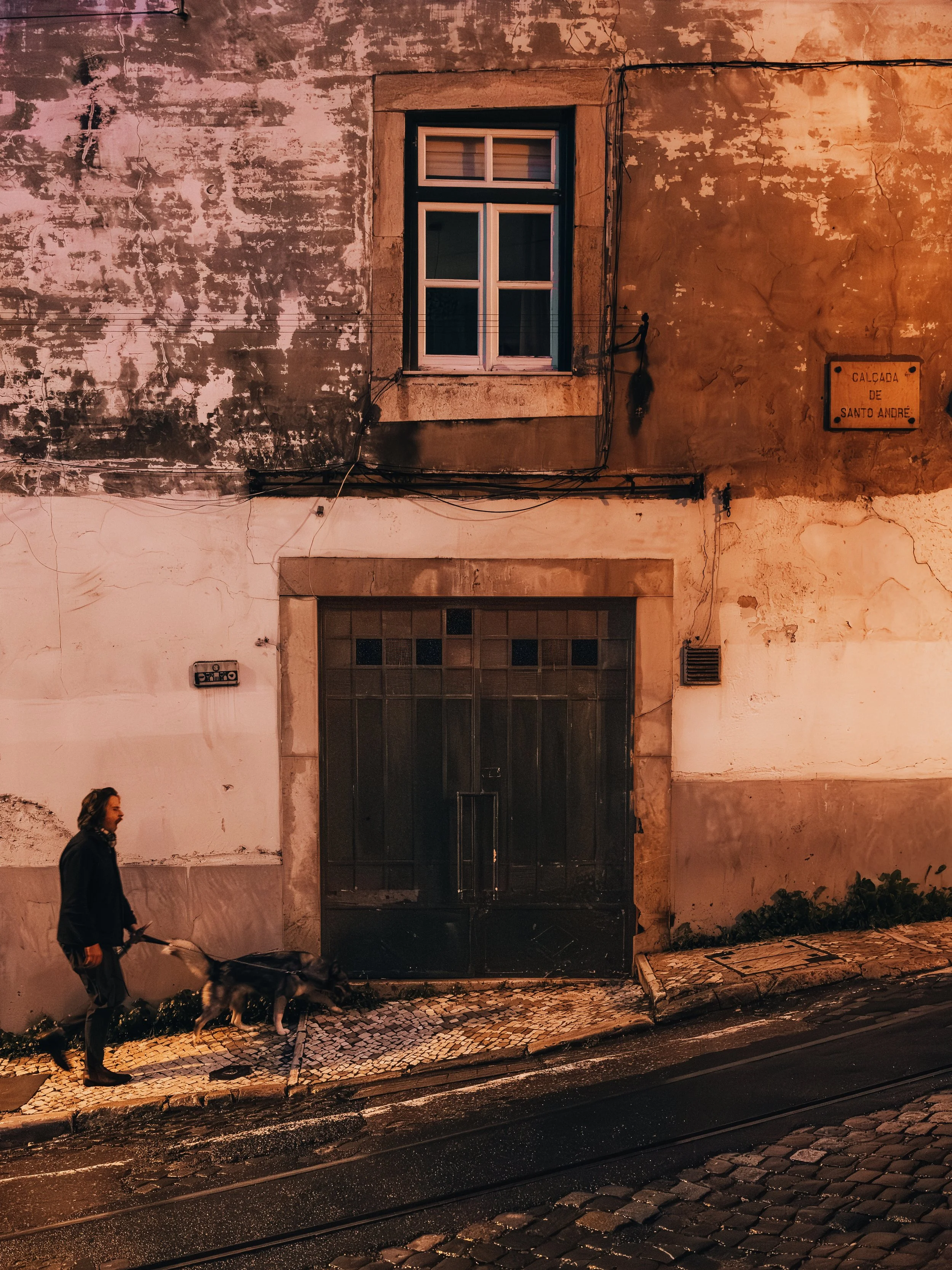 Man wandelt met hond langs een oude, vervallen gebouw op een straat met kasseien.