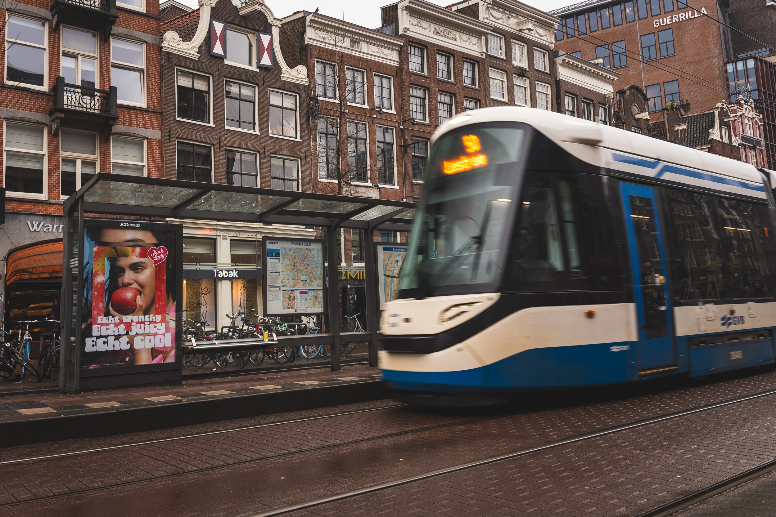 Tram rijdt langs een bushokje met een advertentie in een drukke straat met historische gebouwen in Amsterdam.