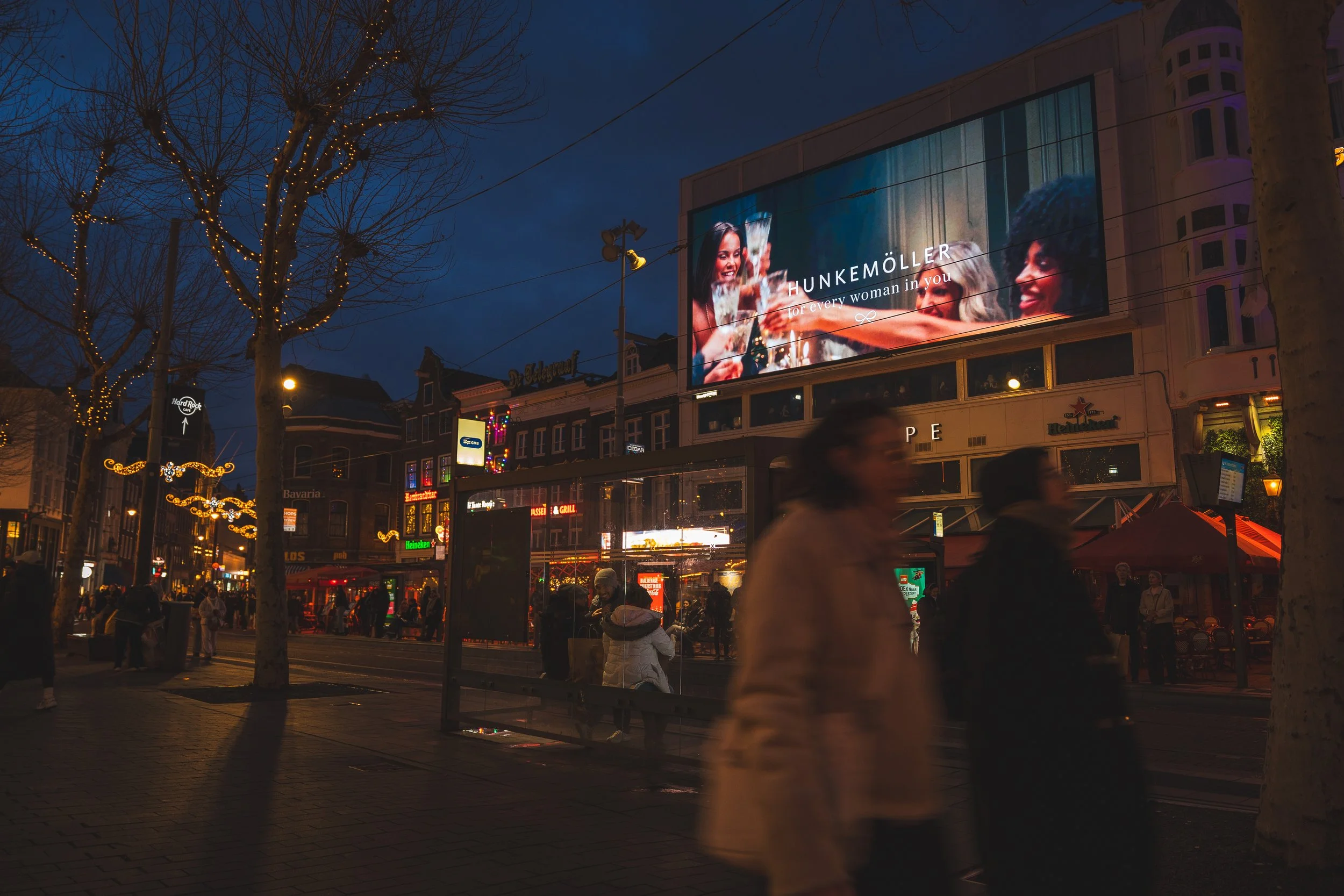 Een levendige stadsstraat in de avond met mensen die wandelen, verlichte bomen en grote digitale billboards die reclame maken.