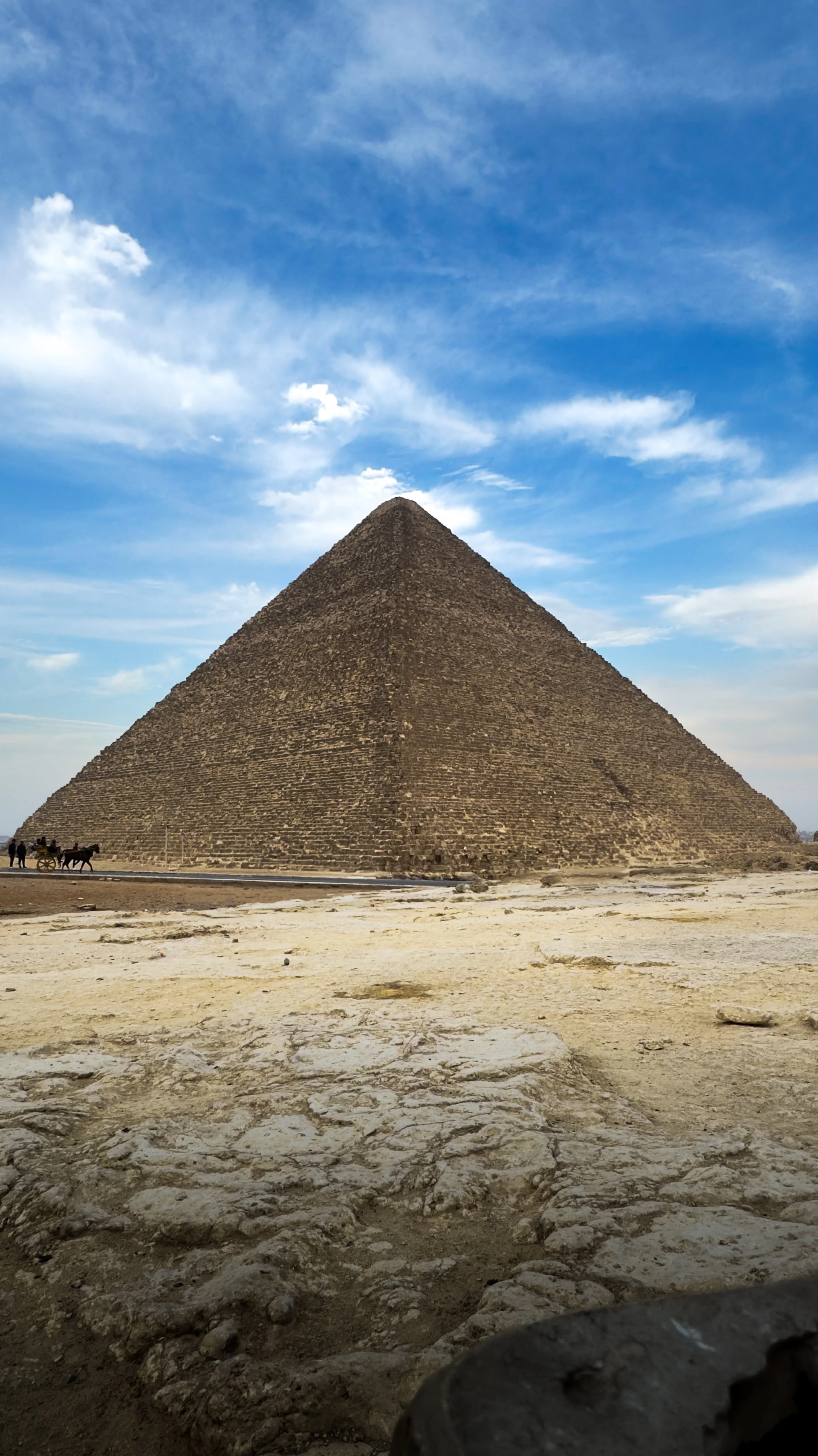 De piramide van Saqqara in Egypte onder een blauwe hemel met wolken, met enkele kamelen en mensen op de voorgrond.