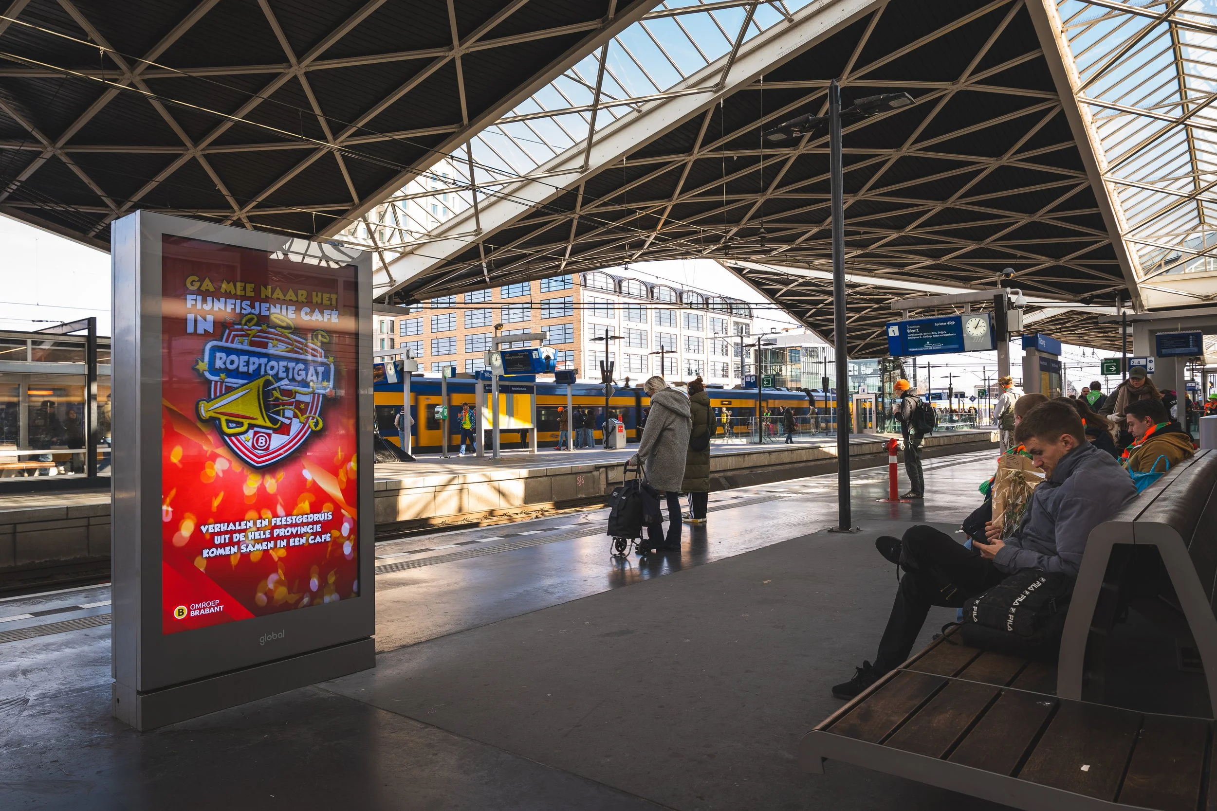 Binnen een druk treinstation zitten reizigers op bankjes, enkele mensen wachten op de trein, een persoon met een trolley en reizigers op het perron, met een groot reclamebord in de voorgrond.