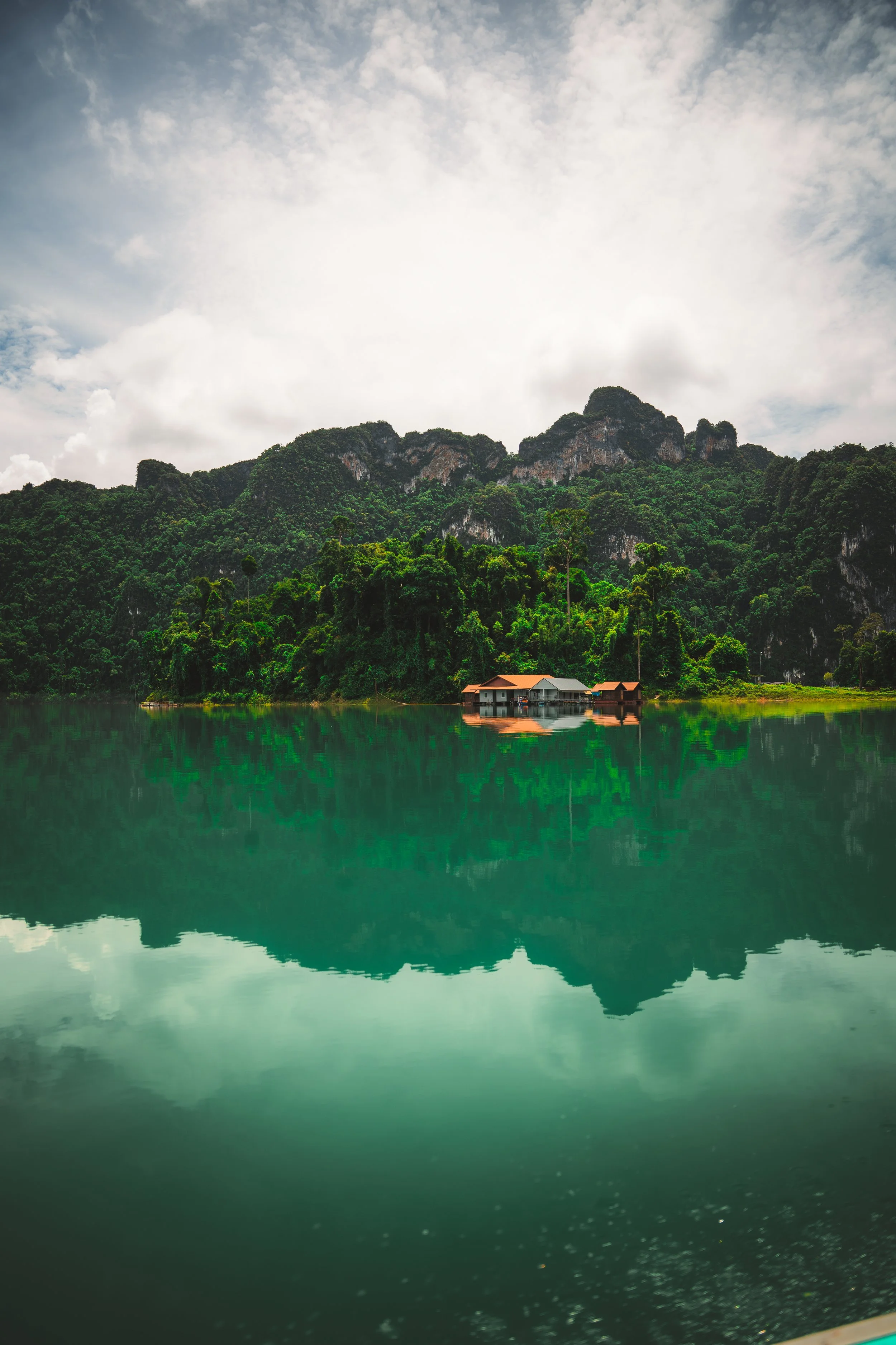 Een huis aan de rand van een meer, omringd door groene bergen en een bewolkte hemel.