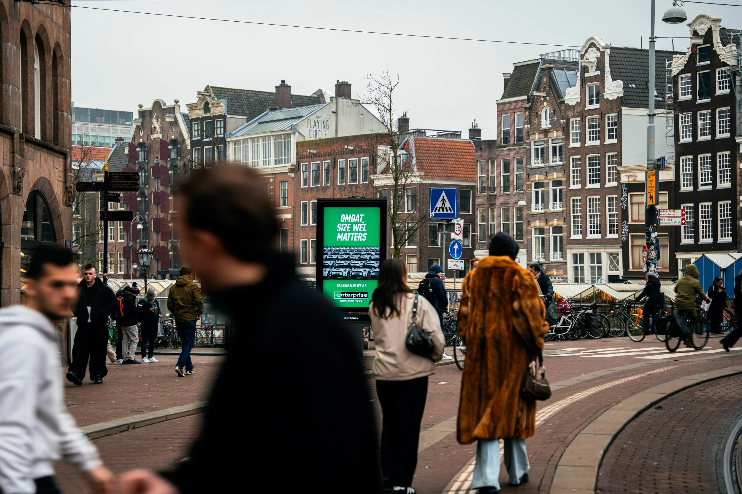 Straatbeeld in een drukke stad met mensen die wandelen, fietsen en wachten bij een zebrapad, omgeven door historische gevels en een digitale reclamezuil met groene tekst, op een bewolkte dag.