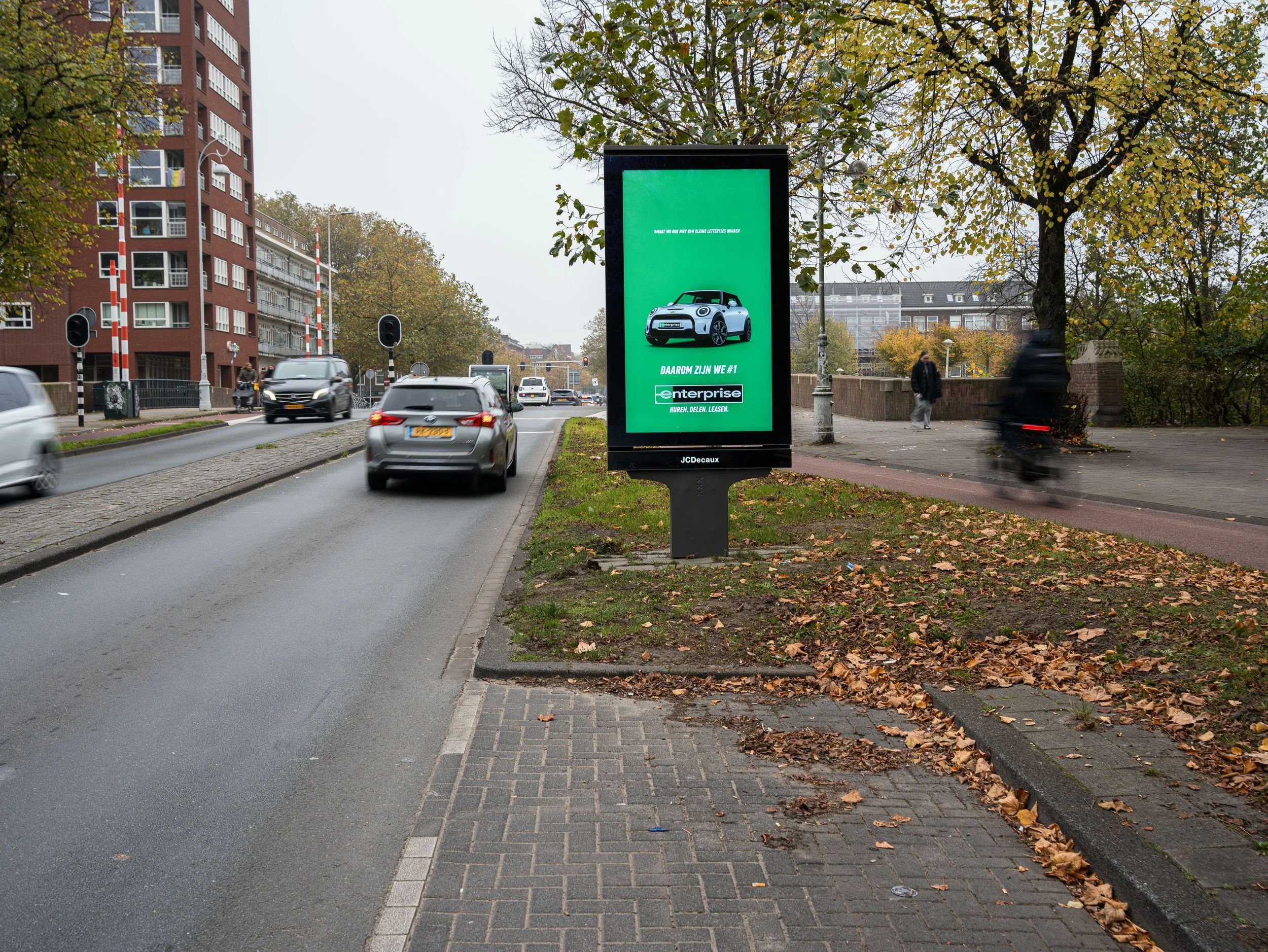Stadstraat met auto verkeer, een digitale reclamezuil langs de stoep met een advertentie voor een auto, bomen met herfstbladeren, enkele voetgangers, en een véloër die voorbijrijdt.